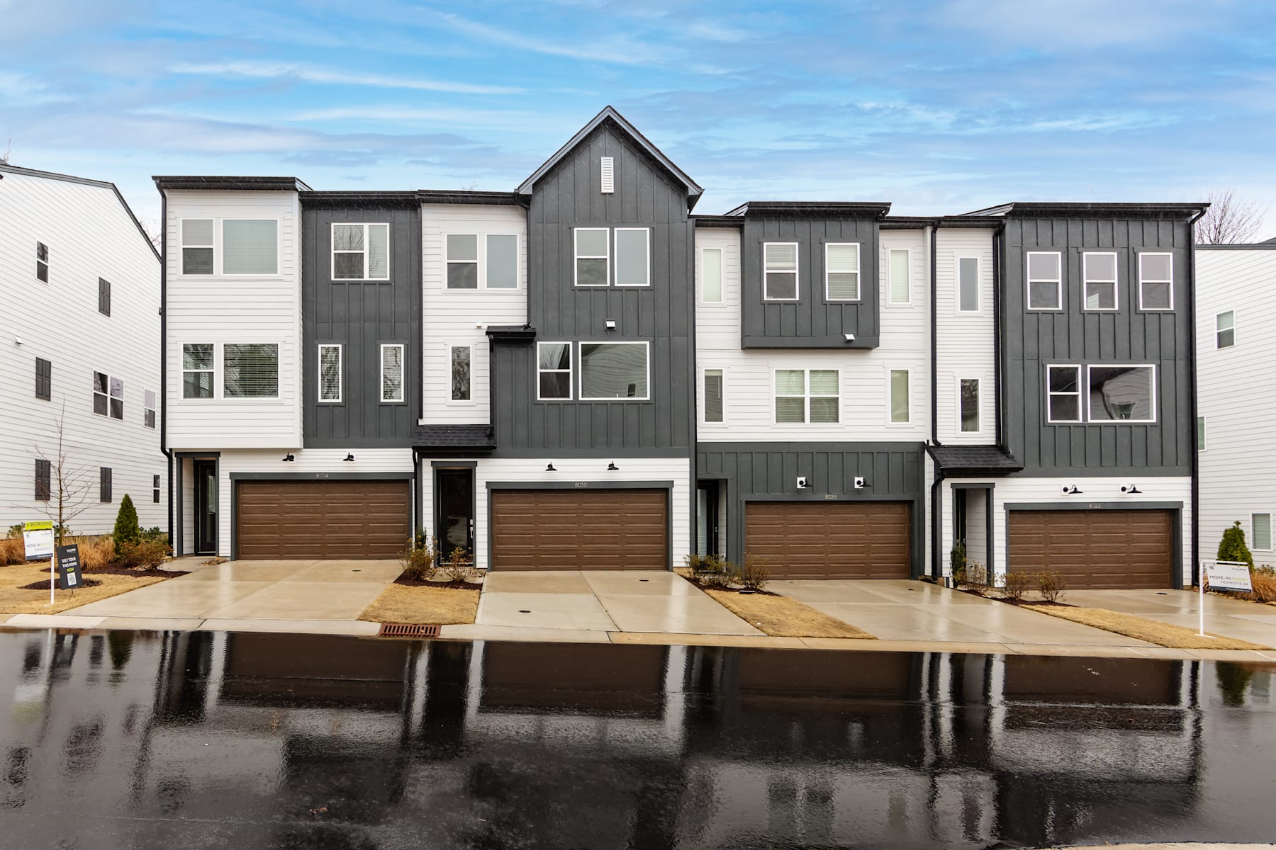 A row of modern, multi-story townhouses with gray and white exteriors, surrounded by a paved area with puddles reflecting the cloudy sky above.