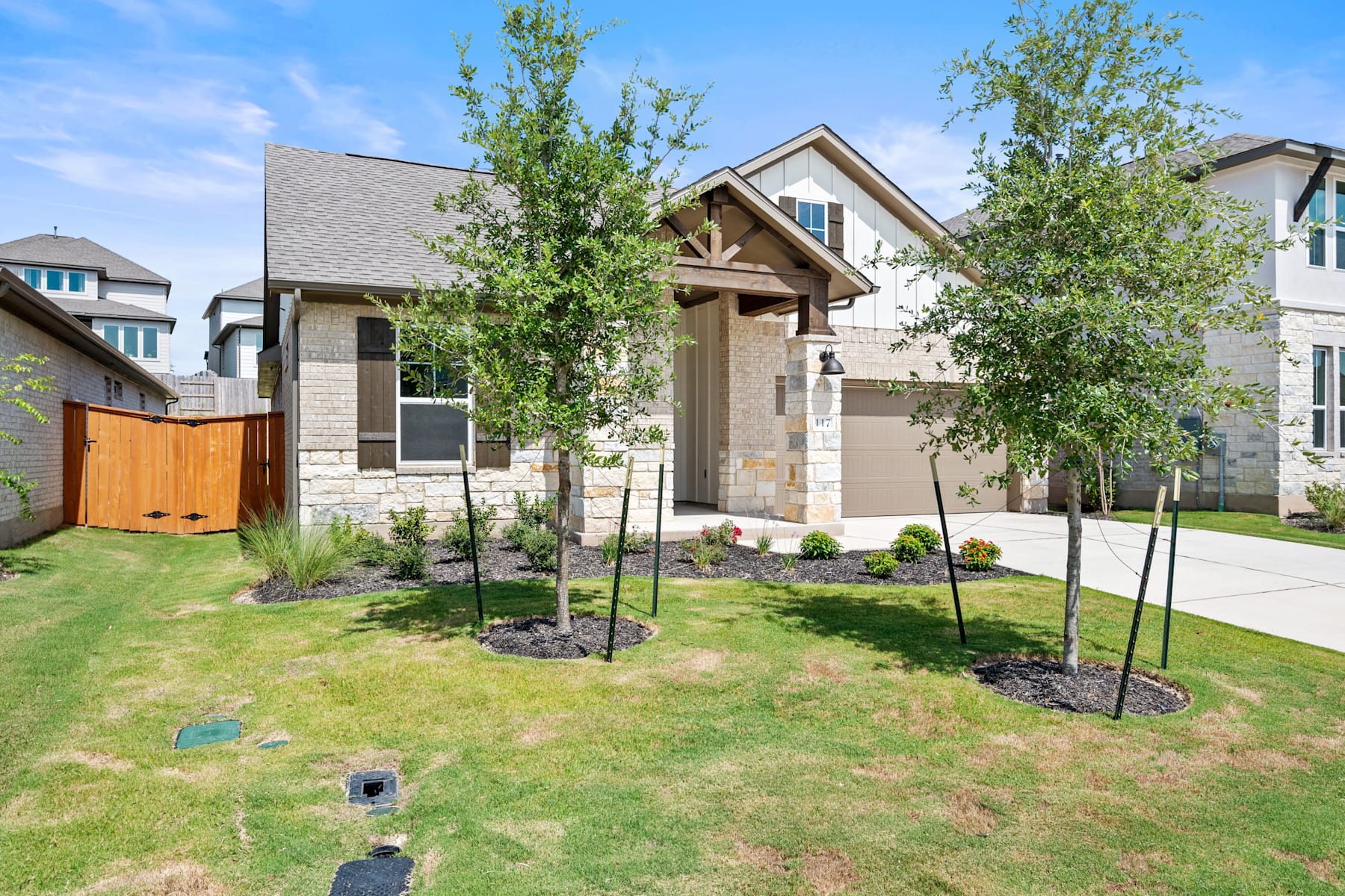 A well-manicured lawn surrounds a two-story house with a stone exterior, a covered porch, and lush greenery in the background.