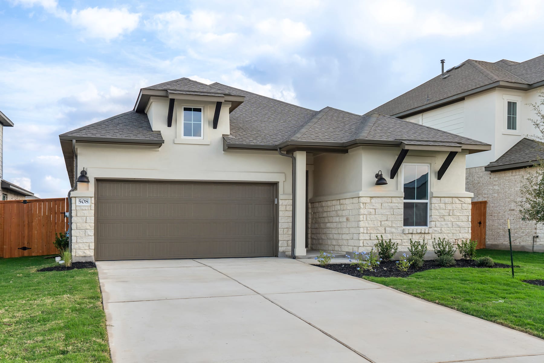 A two-story residential house with a garage, surrounded by a grassy lawn and a wooden fence in the foreground, set against a cloudy sky.