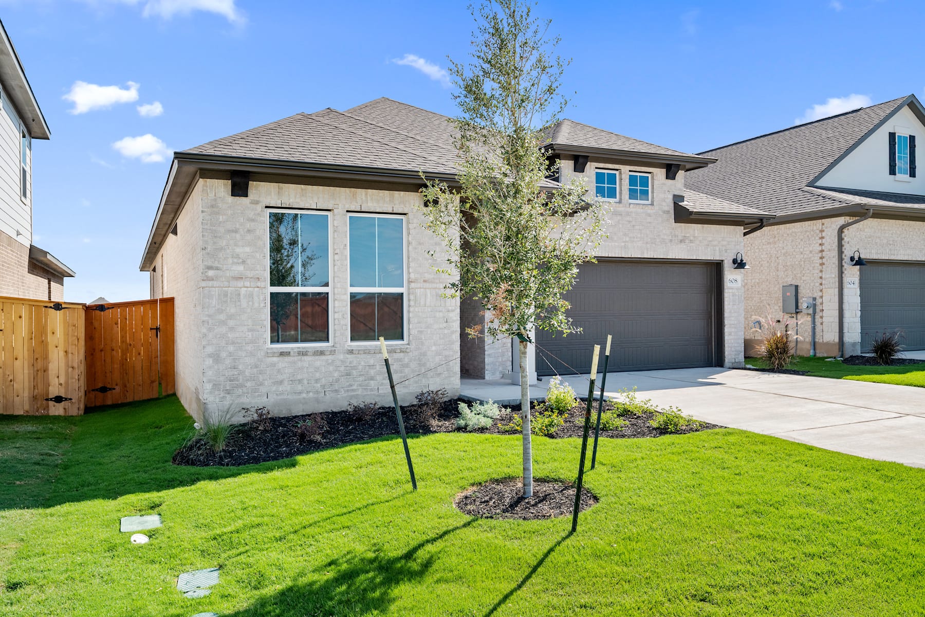 A well-manicured lawn surrounds a modern two-story house with a gray exterior, a tiled roof, and large windows, set against a clear blue sky with scattered clouds.
