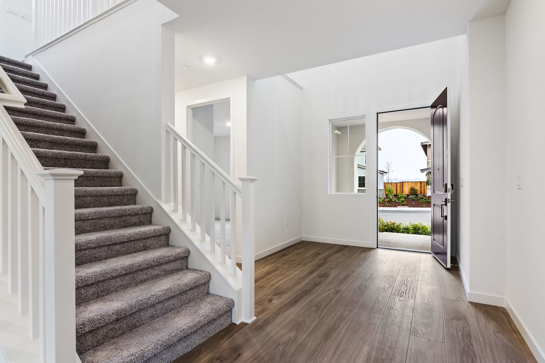 A spacious and well-lit entryway with a carpeted staircase leading up, a wooden floor, and a glass door providing a view of the outdoor landscape.