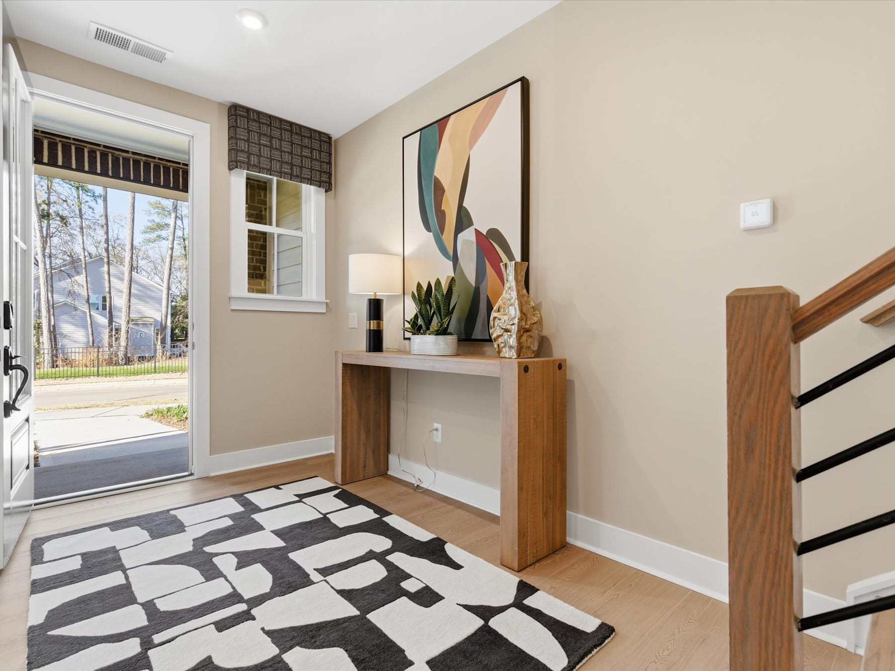 A modern and stylish entryway with a wooden console table, a colorful abstract artwork, and a patterned area rug on the floor, set against a neutral-toned wall and a glass door leading to the outdoors.