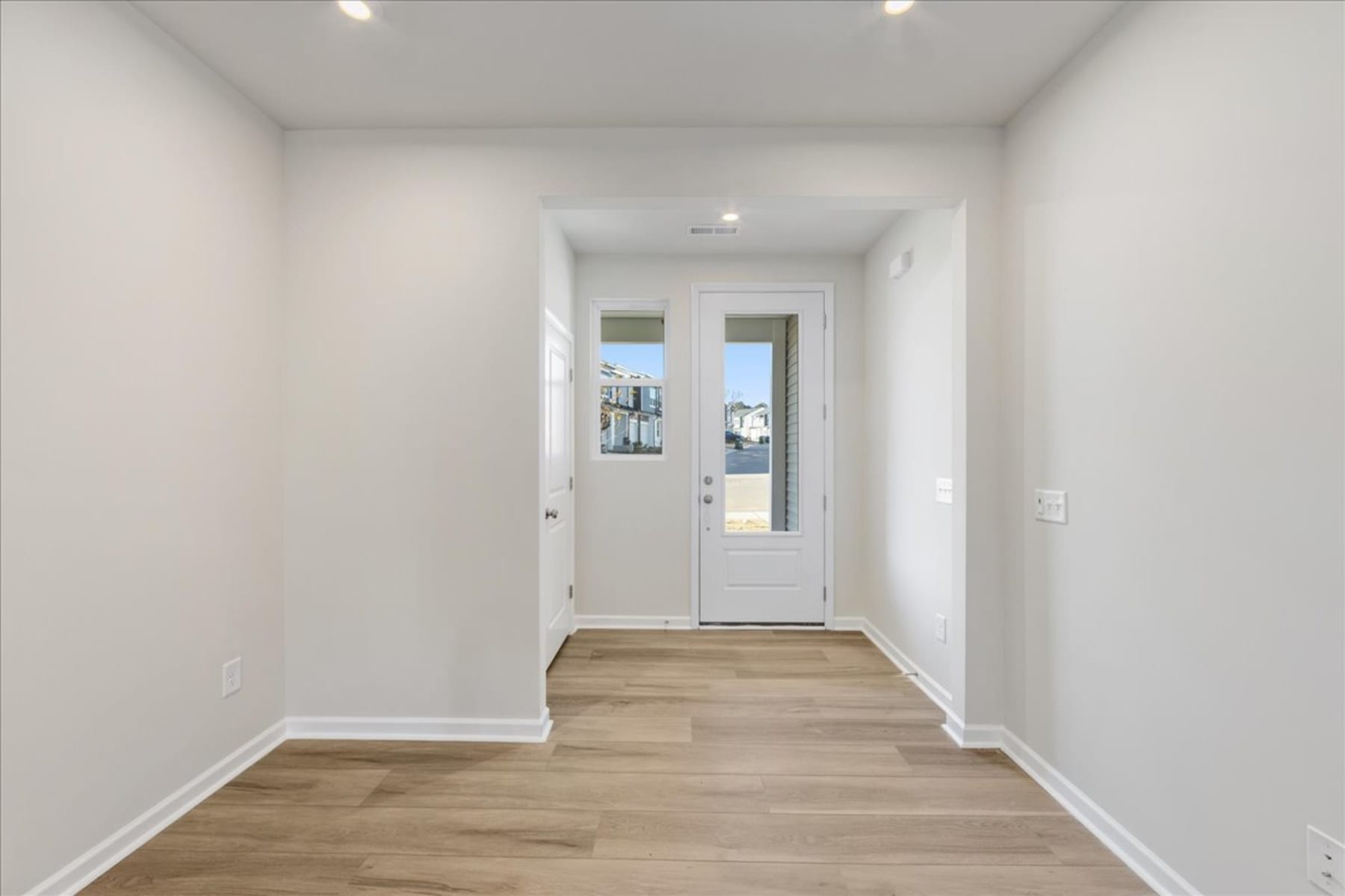 A bright, minimalist hallway with a wooden floor and a white door leading to the exterior.