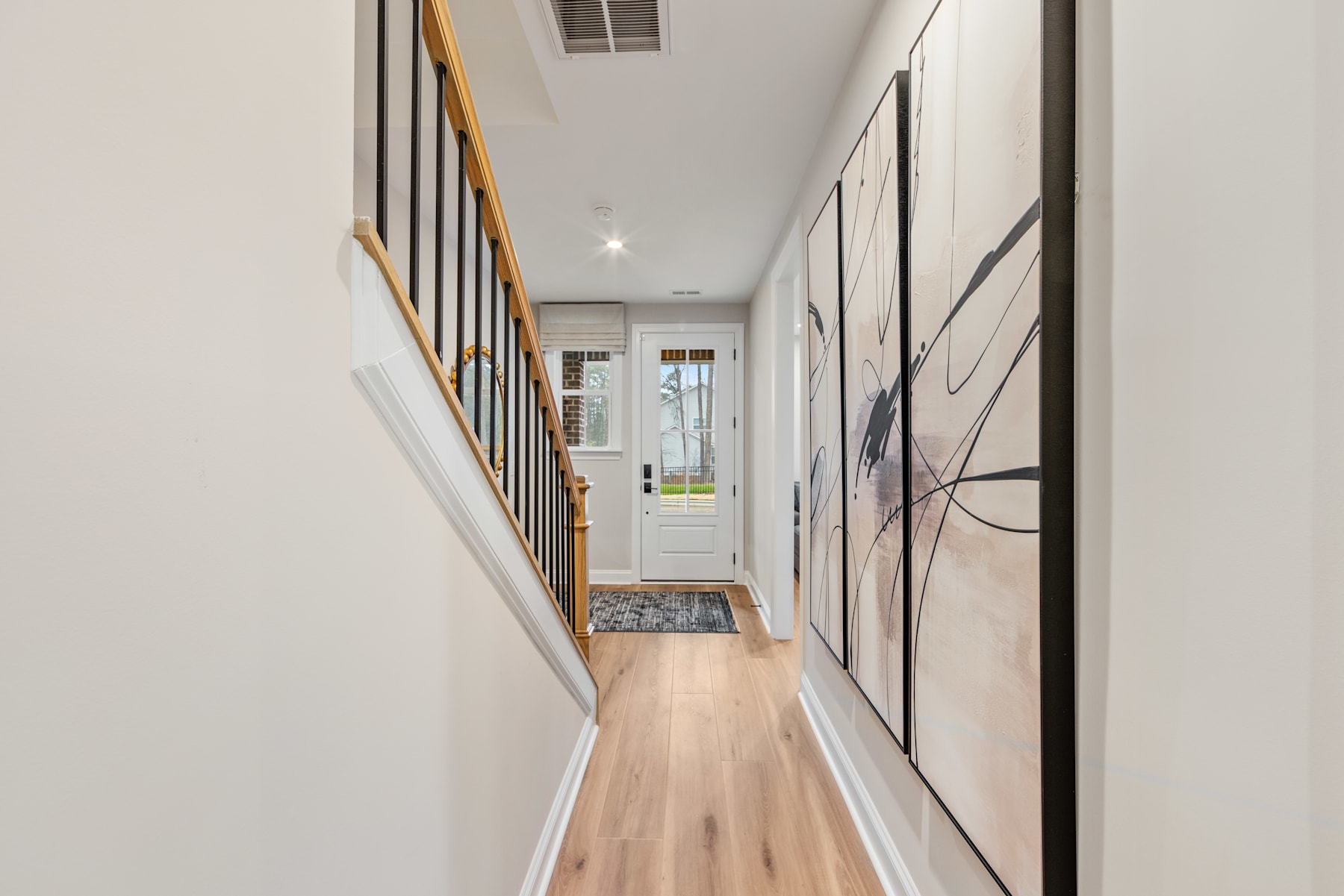 A long, narrow hallway with white walls, wooden floors, and a glass door leading to the outdoors at the far end. The hallway features a wooden railing and several framed artworks or photographs on the walls.