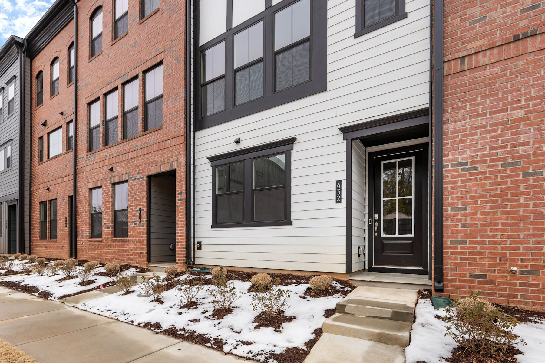 A brick and white building with a snow-covered landscape in the foreground, featuring a wooden door and steps leading up to the entrance.