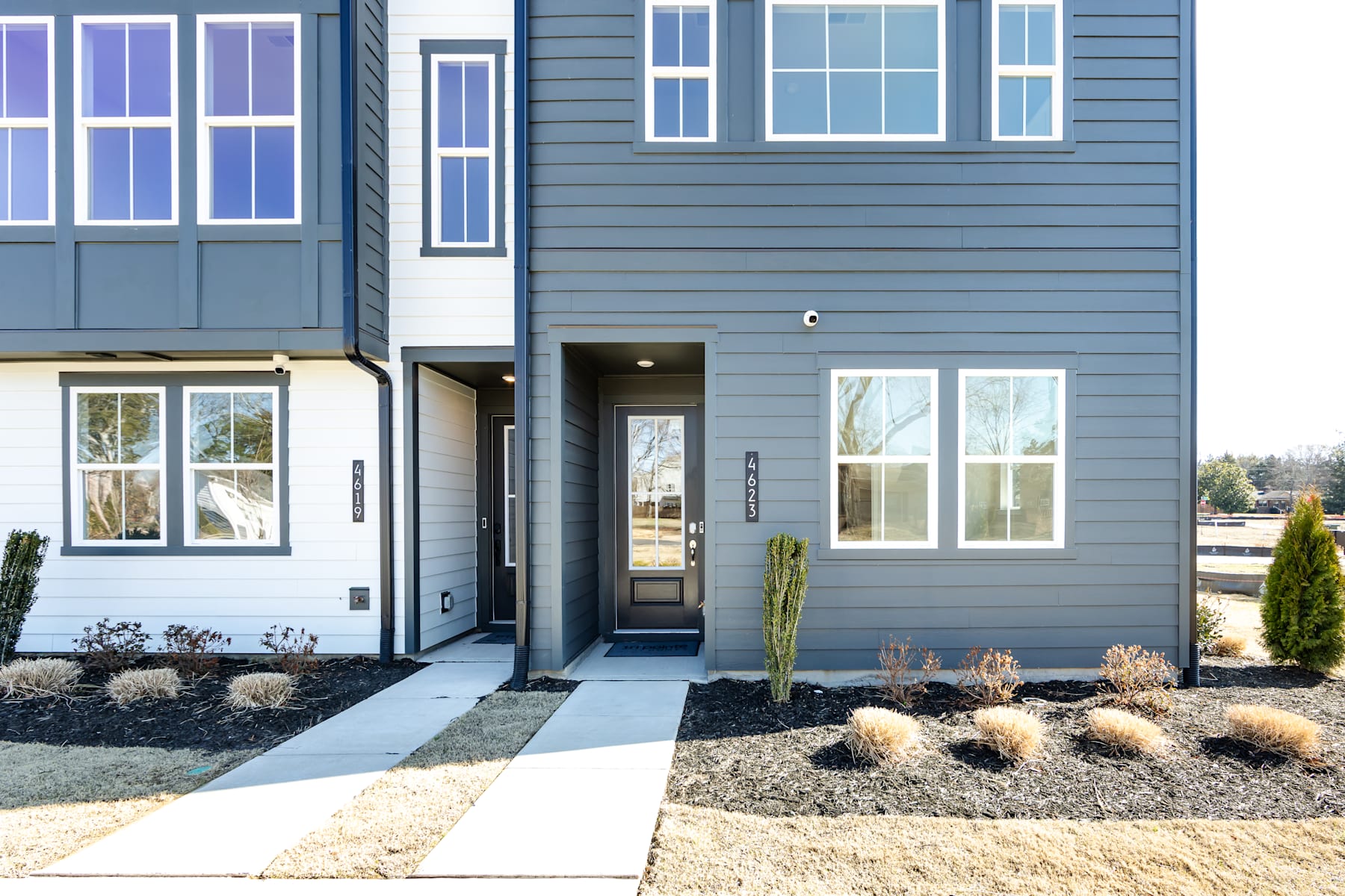 A modern, two-story residential building with a gray exterior, featuring large windows, a front door, and a landscaped yard with gravel and cacti in the foreground.
