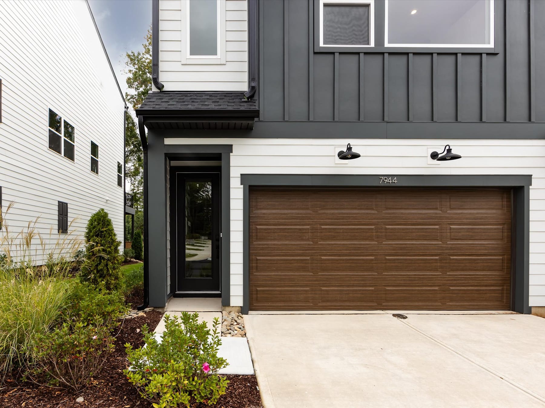 A modern, two-story house with a wooden garage door, surrounded by landscaped greenery and a concrete driveway.