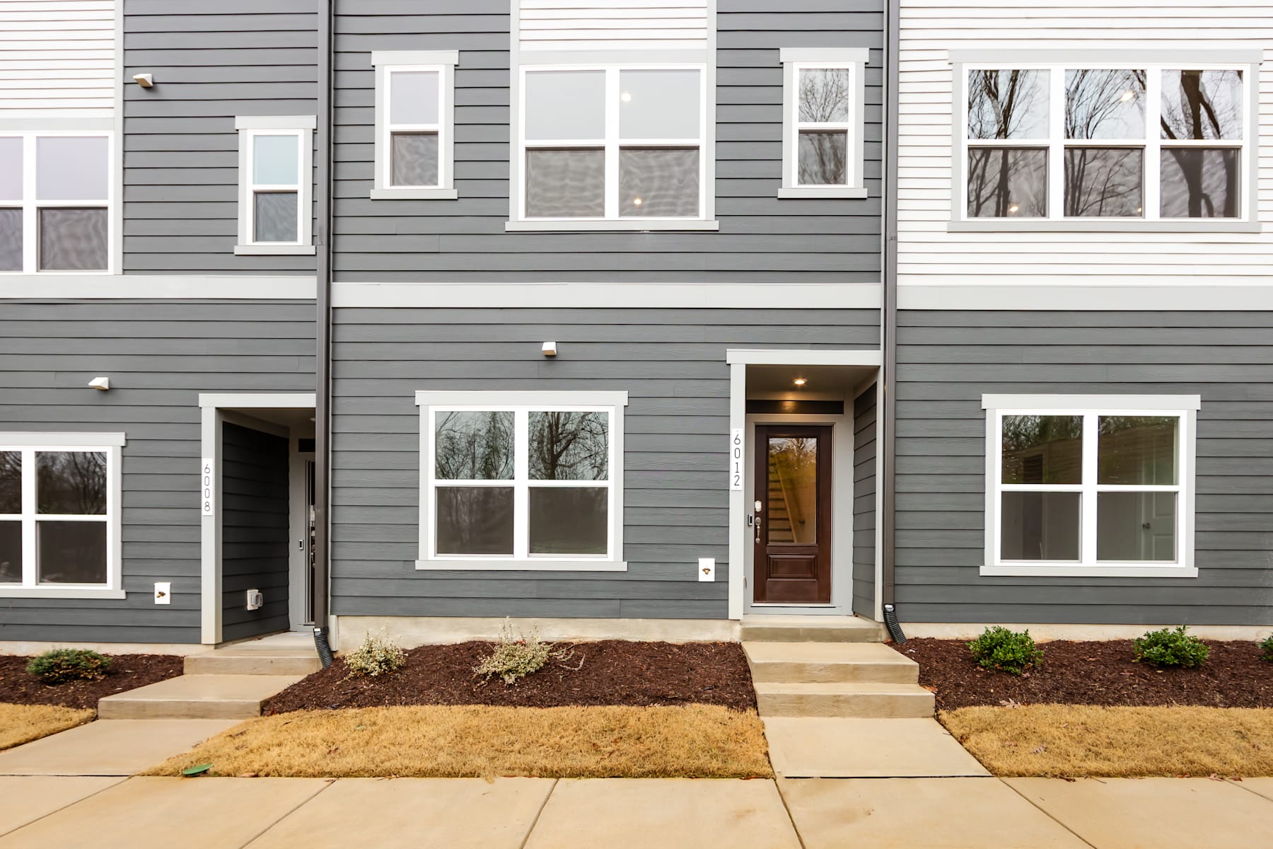 A modern townhouse with a gray exterior, featuring a paved walkway leading to the front door, surrounded by landscaped beds with plants.