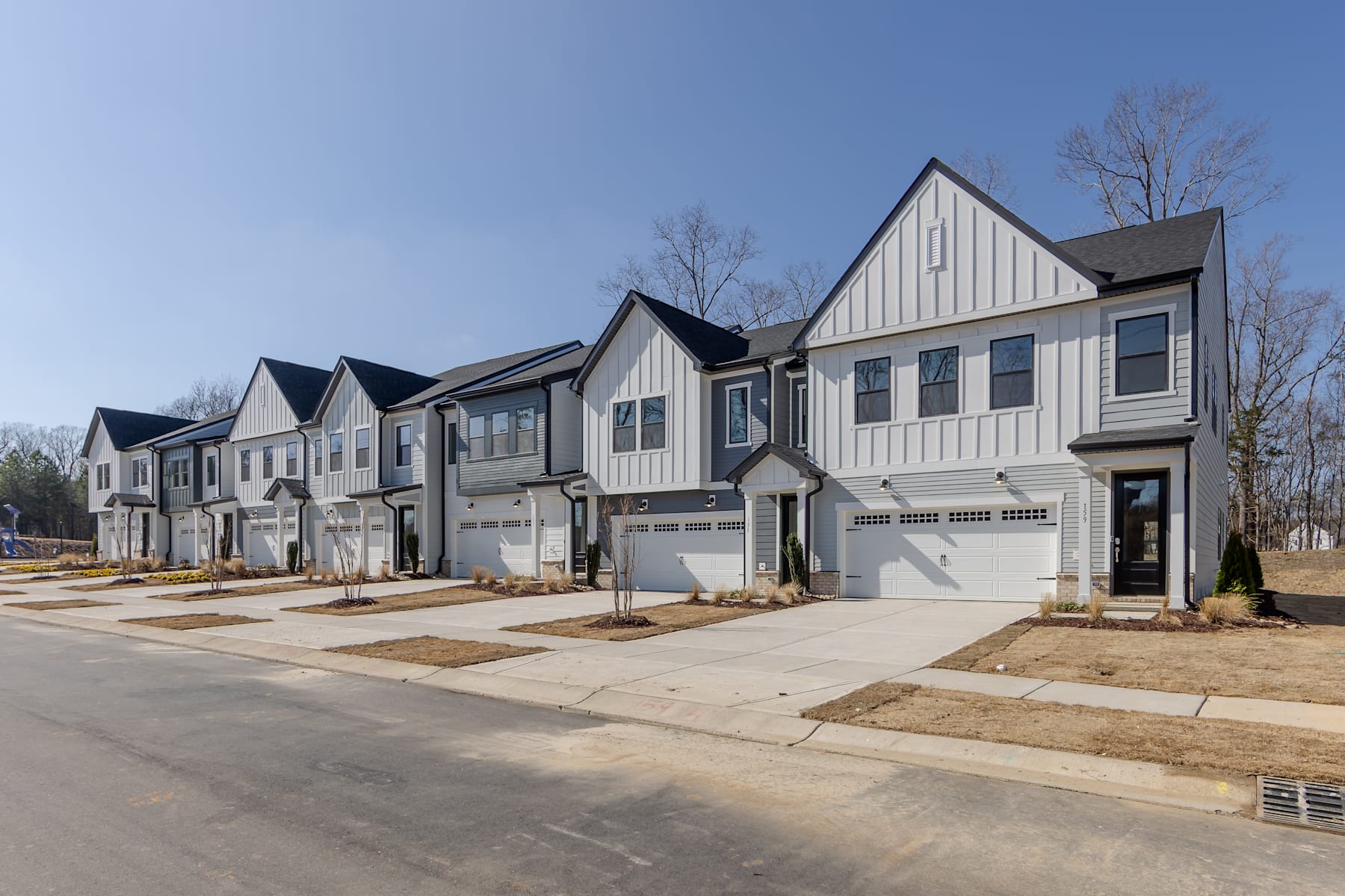A row of modern, two-story townhouses with white siding and black roofs, set against a clear blue sky and surrounded by trees in the background.