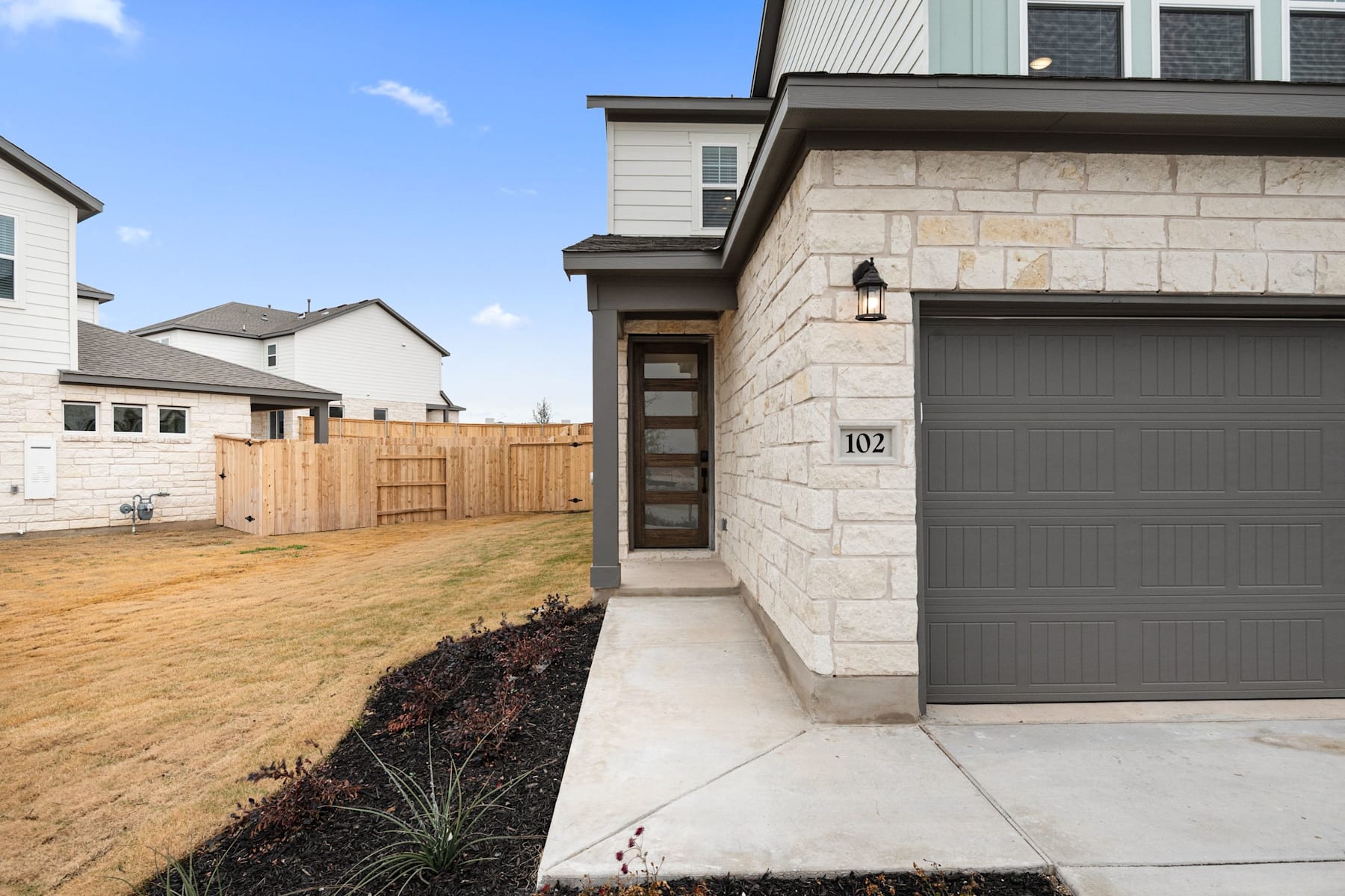 A modern two-story house with a stone exterior, a gray garage door, and a wooden fence in the foreground, set against a clear blue sky.