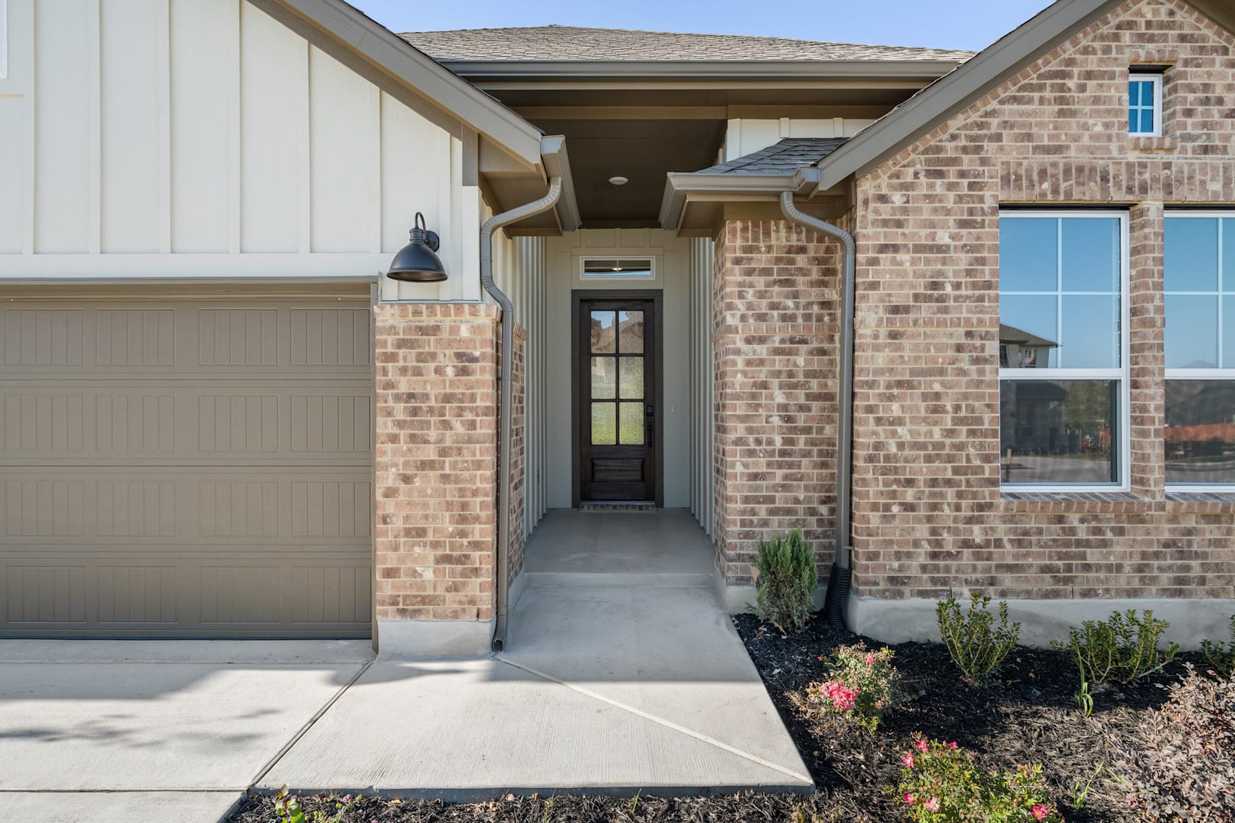 A brick and siding home with a covered entryway, a garage door, and a landscaped front yard with plants and a concrete walkway.