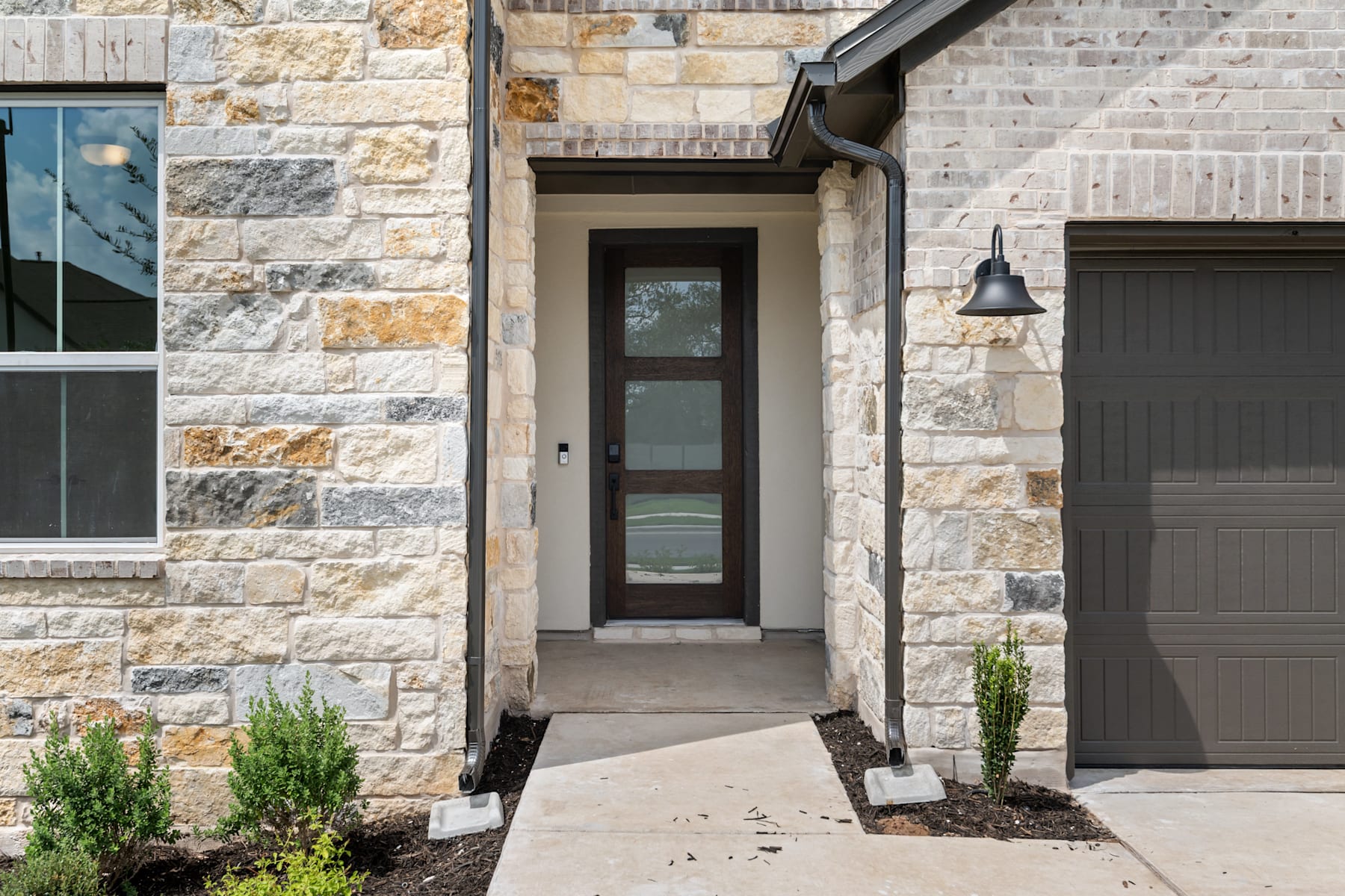 The image shows the entrance to a stone-clad house, with a wooden door and glass panel, surrounded by landscaping elements such as potted plants and a paved walkway leading to the entrance.