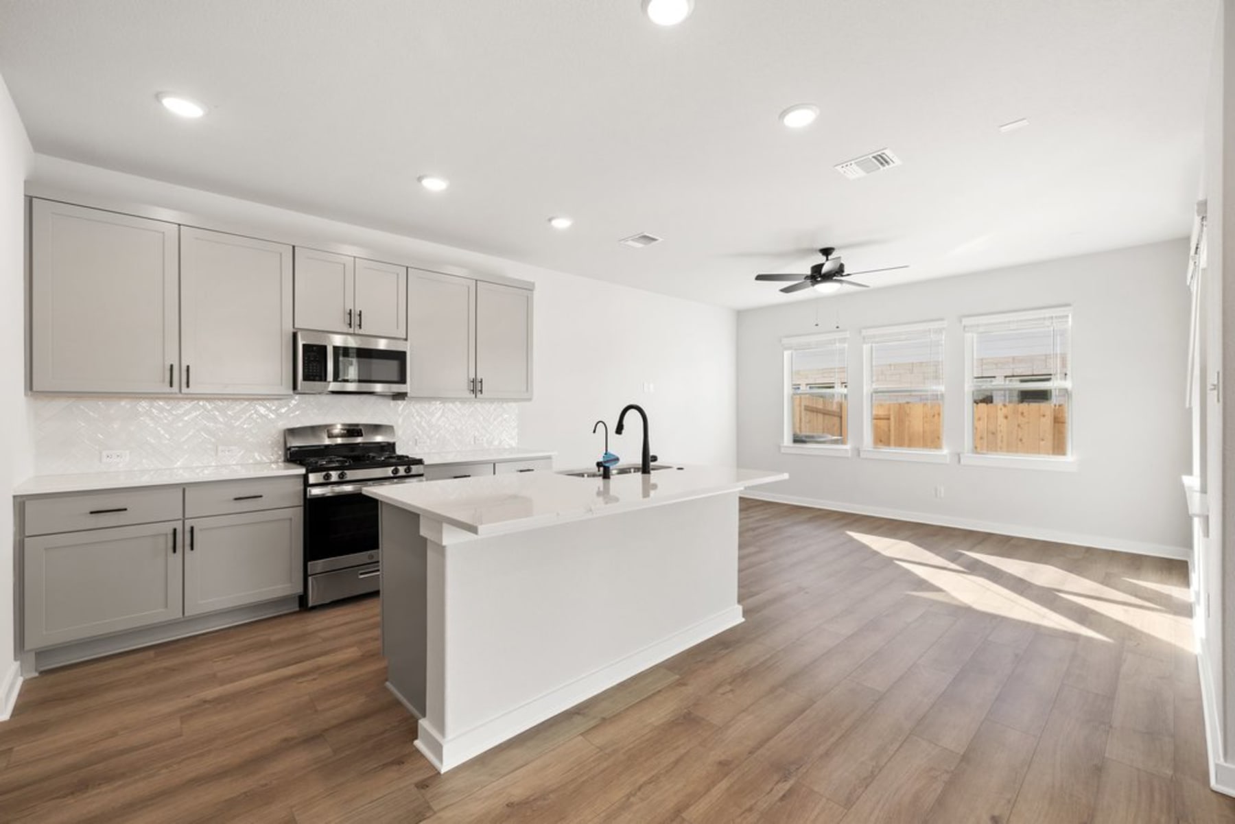 A modern, open-concept kitchen with gray cabinets, stainless steel appliances, and a white countertop island, set against a bright, airy living space with hardwood floors.