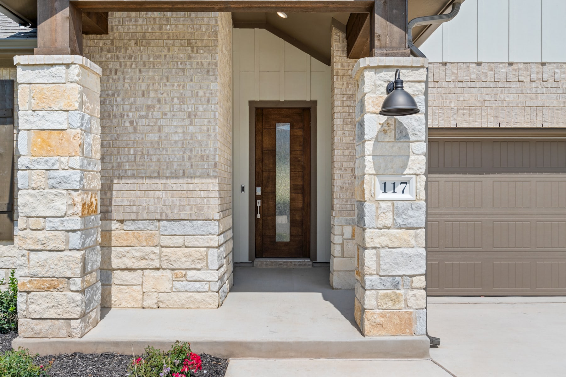 The image shows the exterior of a modern, stone-clad house with a wooden front door, a garage door, and a porch light fixture.
