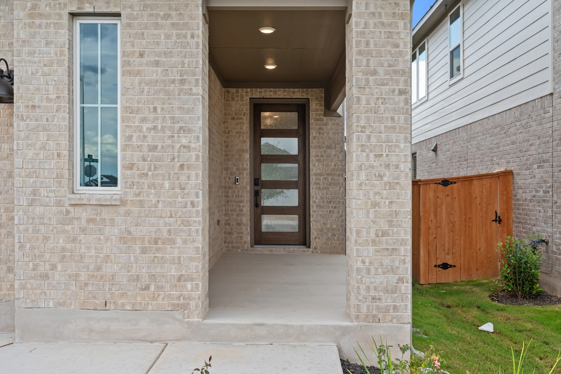 A brick-walled entryway with a wooden door and glass panels leads to a grassy yard with a wooden fence in the background.