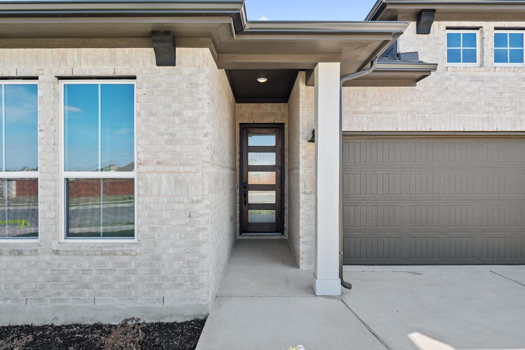 The image shows a modern, two-story residential building with a garage door, large windows, and a covered entryway leading to the front door.