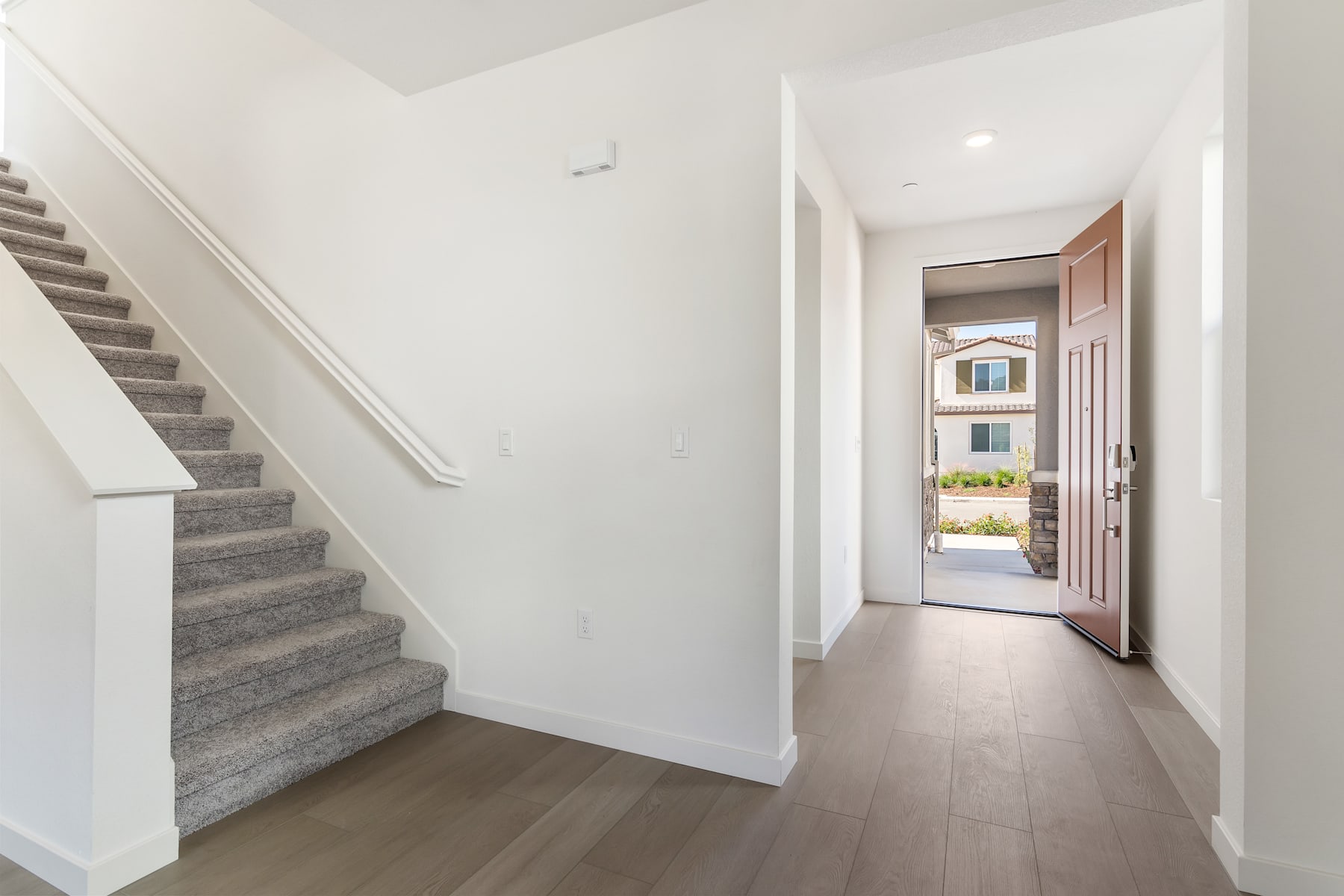 A bright and spacious hallway with a staircase leading upstairs, and a doorway visible in the background, revealing a glimpse of the exterior.