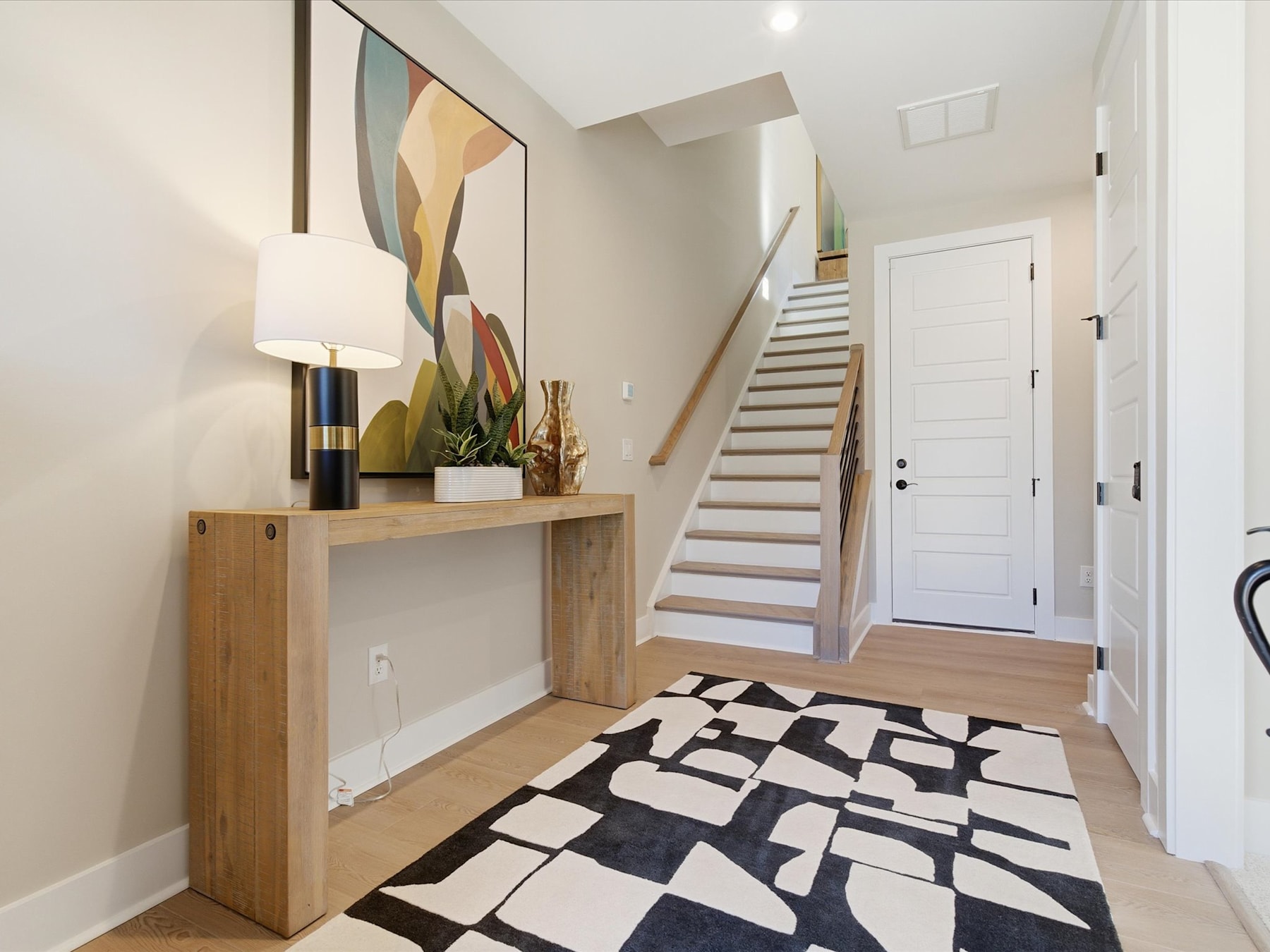 A modern and minimalist entryway with a wooden console table, a decorative vase, and a patterned rug leading up to a staircase.