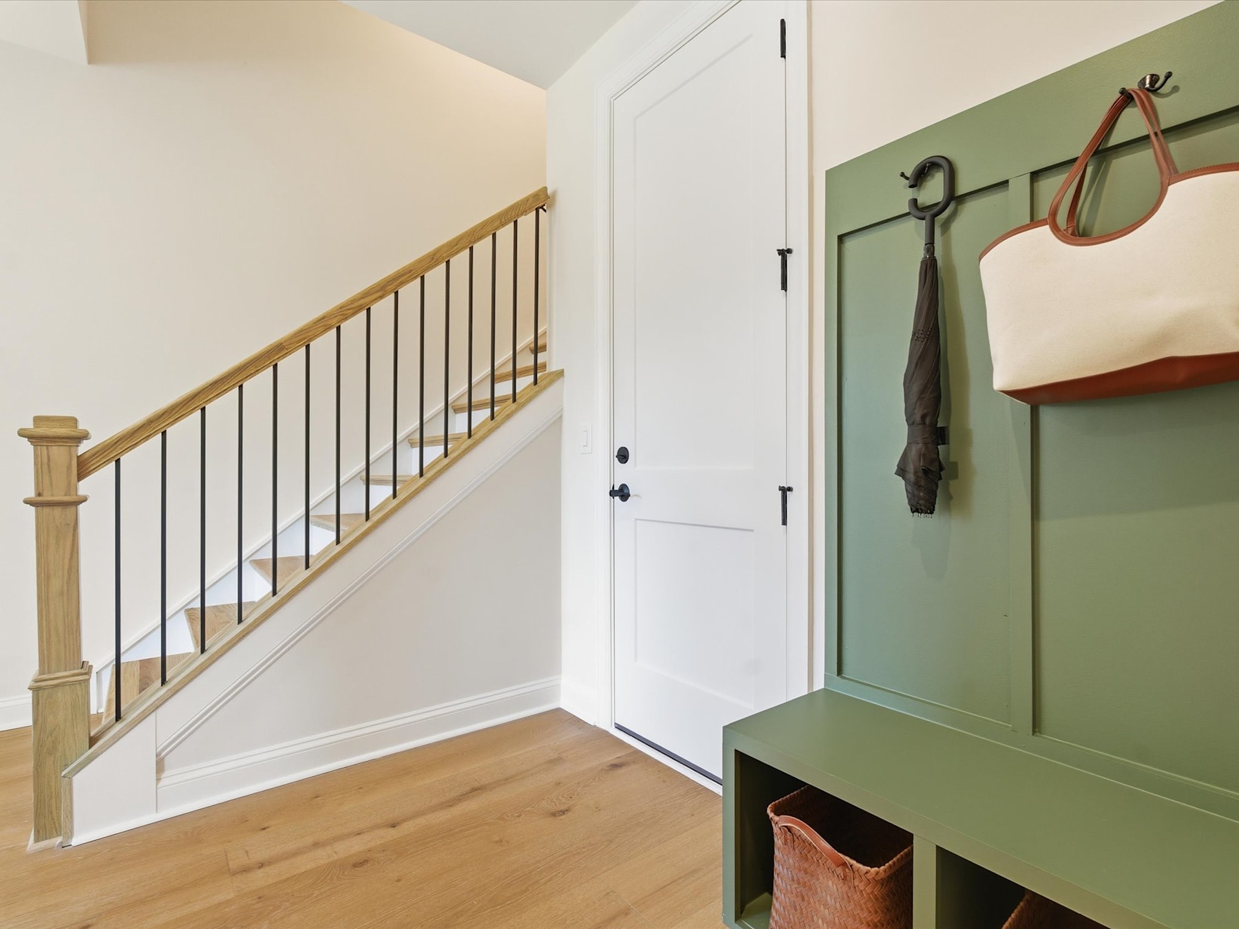 A cozy entryway with a wooden staircase, a white door, and a green storage bench with hanging hooks, set against a neutral-toned wall.