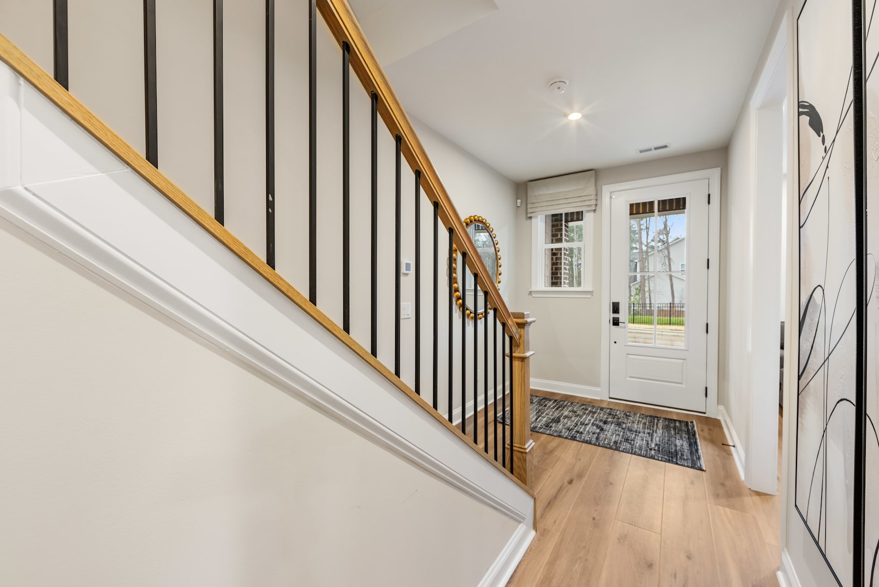 A bright and airy hallway with a wooden staircase, white walls, and a patterned rug leading to a glass-paneled door that provides a view of the outdoors.