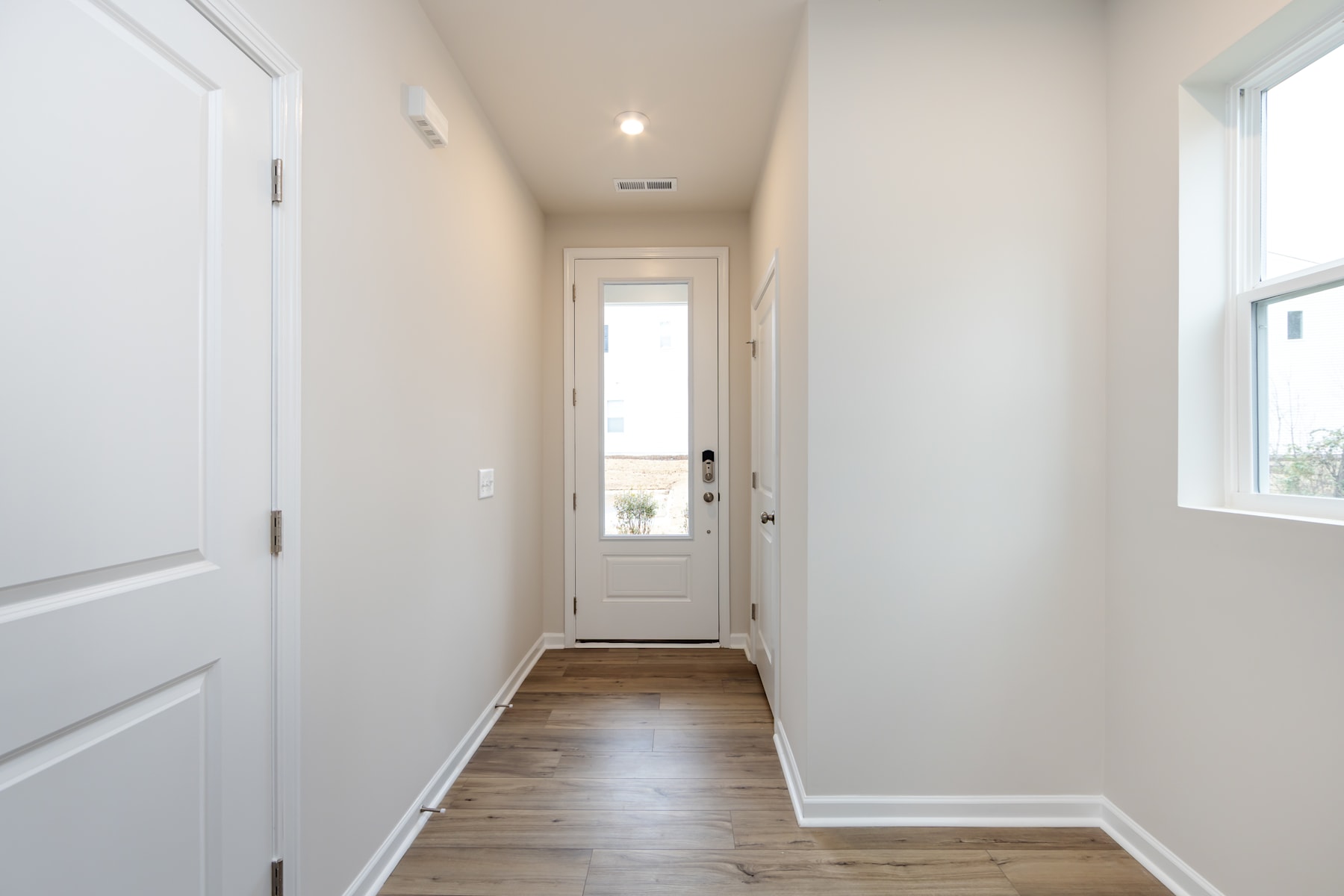 A bright, airy hallway with white walls, a wooden floor, and a glass-paneled door leading to the exterior.