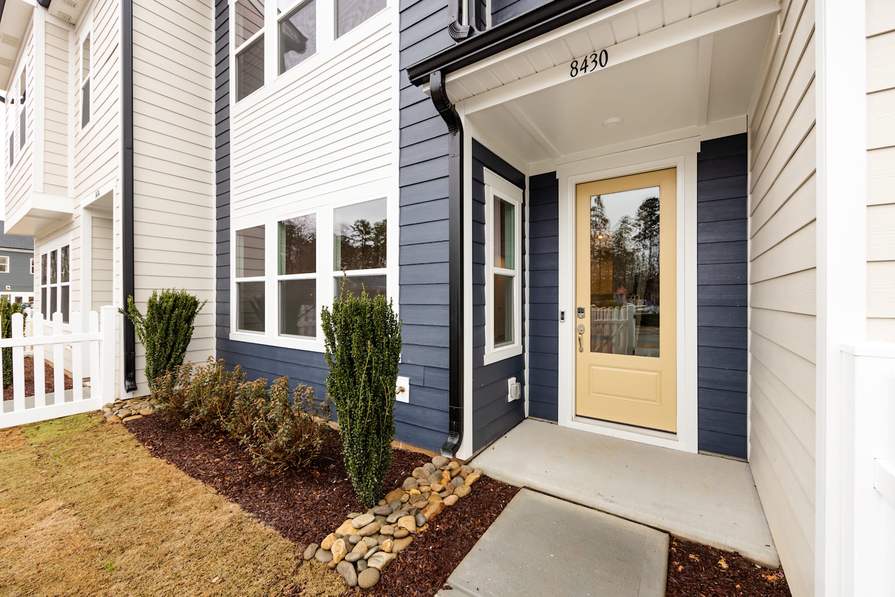 The image shows a modern, two-story residential building with a porch and a landscaped front yard featuring a stone pathway leading to the entrance.