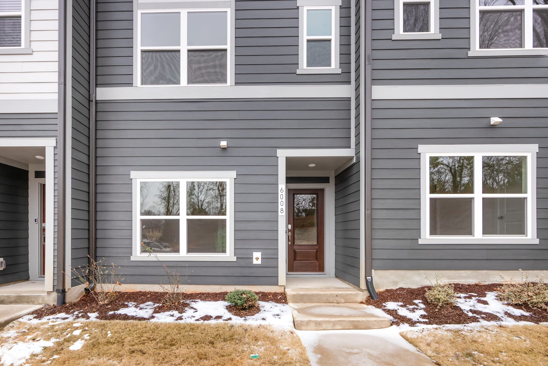 A two-story gray-sided townhouse with a covered porch, surrounded by a snowy landscape and landscaping in the foreground.