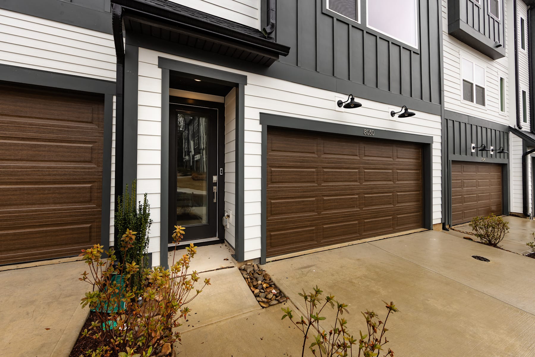 The image shows a modern, multi-story residential building with wooden garage doors and a paved walkway leading to the entrance, surrounded by small plants and shrubs.