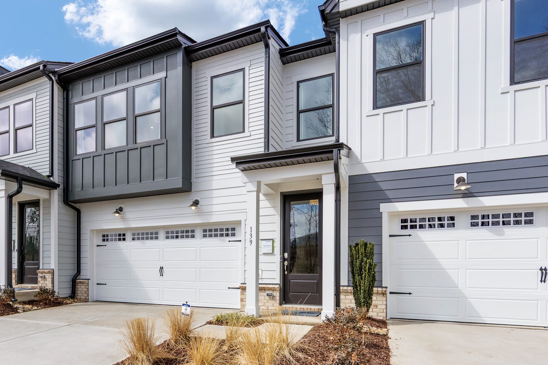 The image depicts a modern townhouse complex with gray siding, white trim, and garage doors. The foreground features a paved driveway and landscaping with small plants, while the background shows a clear sky with some clouds.
