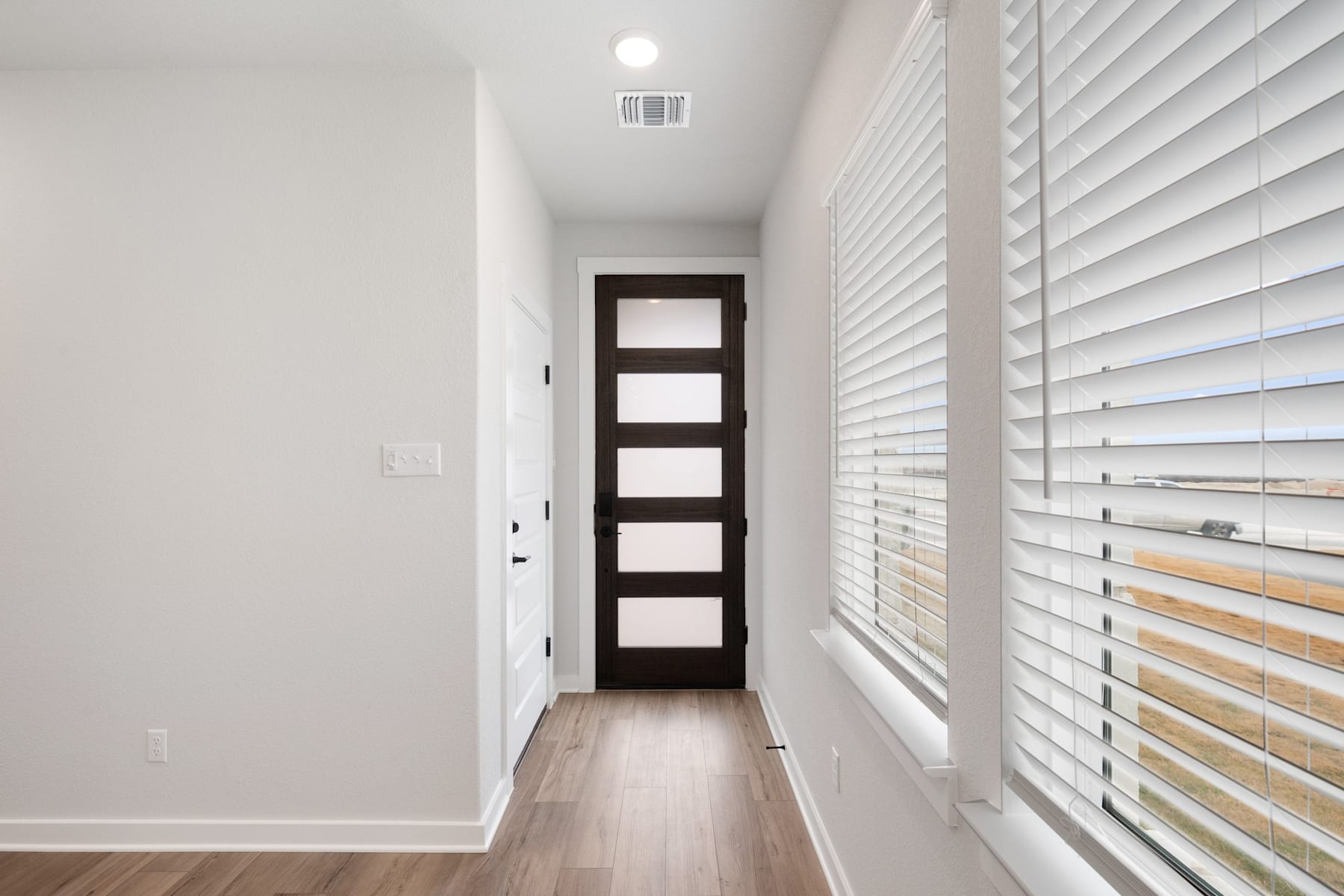 A well-lit, minimalist hallway with a dark wooden door and white walls, complemented by horizontal blinds covering the window.