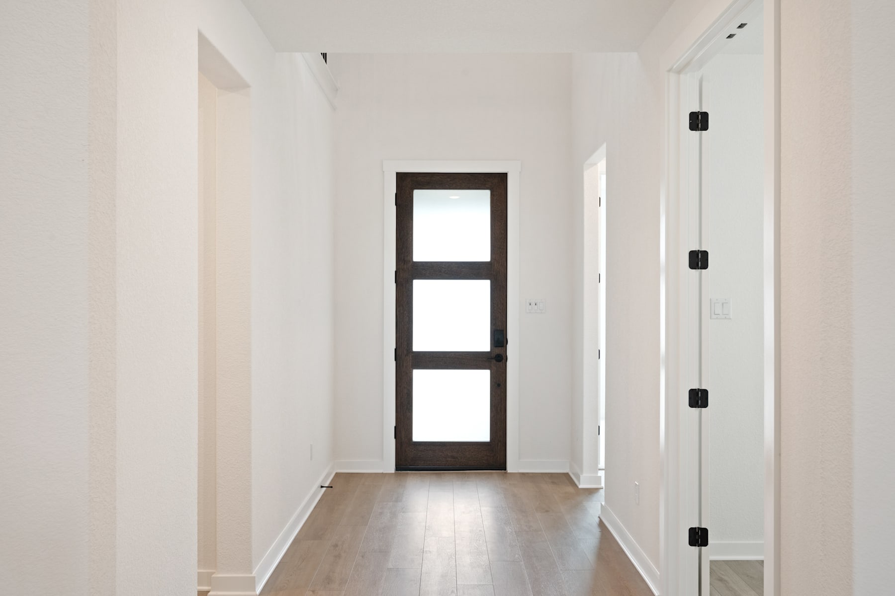 A simple, minimalist hallway with a dark wooden door and a light-colored floor leading to a bright, open space.