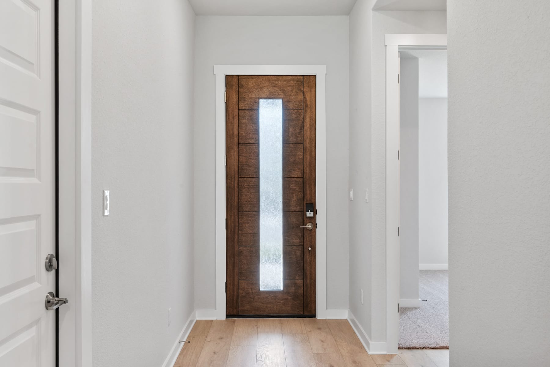 A narrow wooden door with a vertical window panel stands in a bright, white-walled hallway, leading to another room beyond.