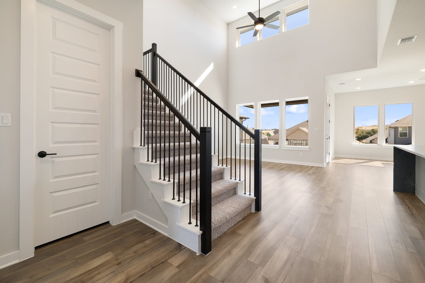 A spacious and bright entryway with a wooden staircase featuring a black metal railing, leading to an open living area with high ceilings and large windows.