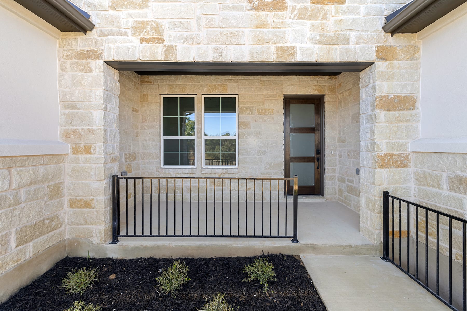 A stone-clad entryway with a metal railing leading to a wooden door, surrounded by a small garden bed with plants.