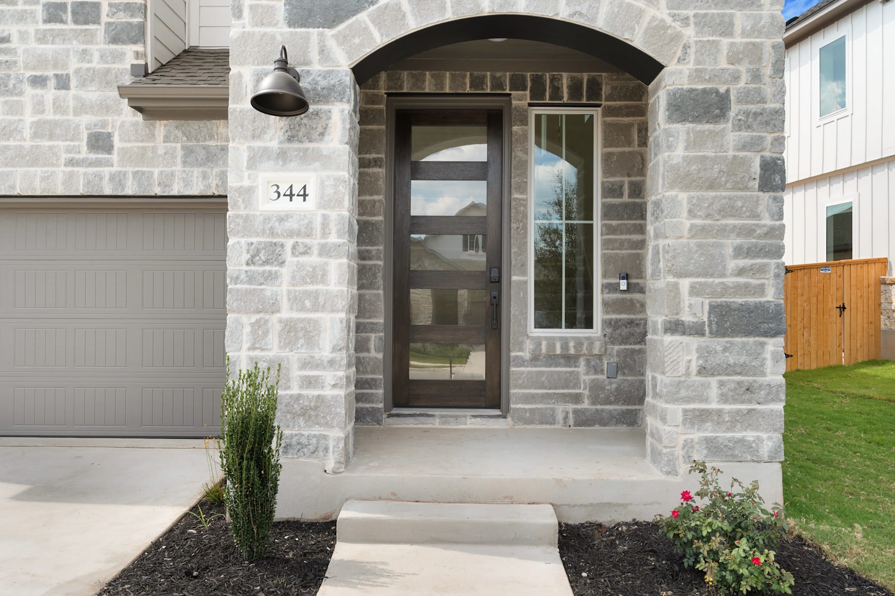 A stone-clad entryway with a wooden door and a garage door, surrounded by landscaping and a grassy yard.
