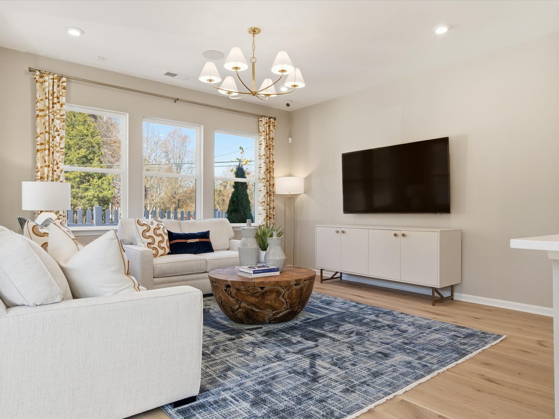 A cozy and well-furnished living room with a large TV, a plush white sofa, a wooden coffee table, and a patterned area rug on a hardwood floor. The room is illuminated by a chandelier and features large windows overlooking a scenic outdoor view.