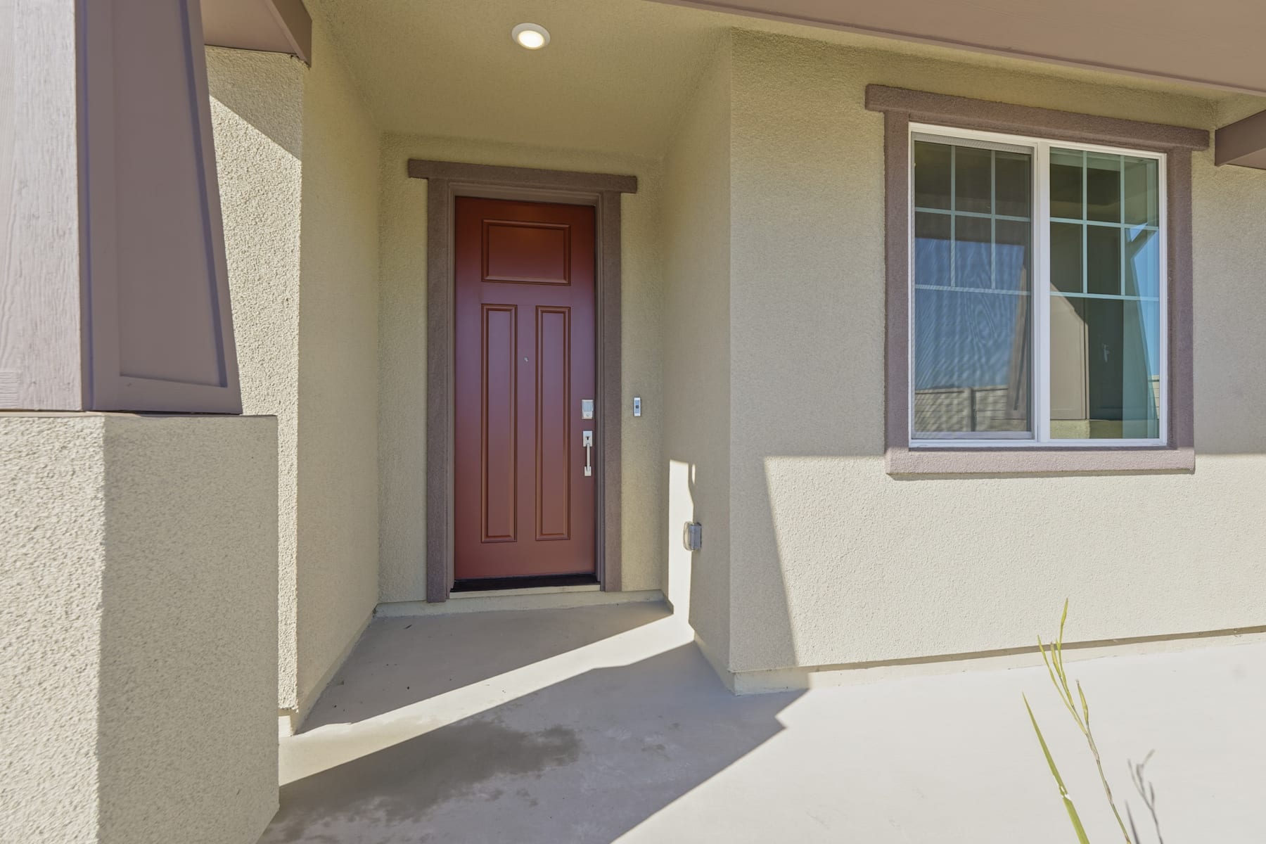 A wooden front door with a glass panel is visible in the foreground, leading to a well-lit and spacious interior. The exterior walls are painted in a light, neutral color, and a window can be seen to the right of the door.
