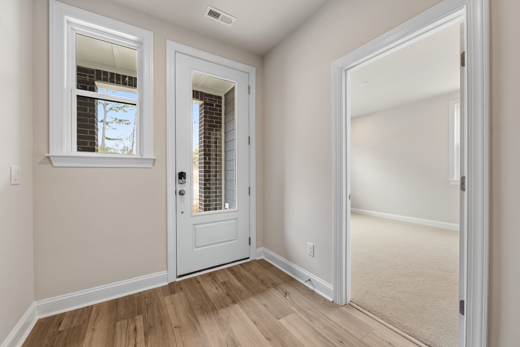 A bright and airy entryway with a white door, windows, and hardwood floors, leading to a hallway and another room.