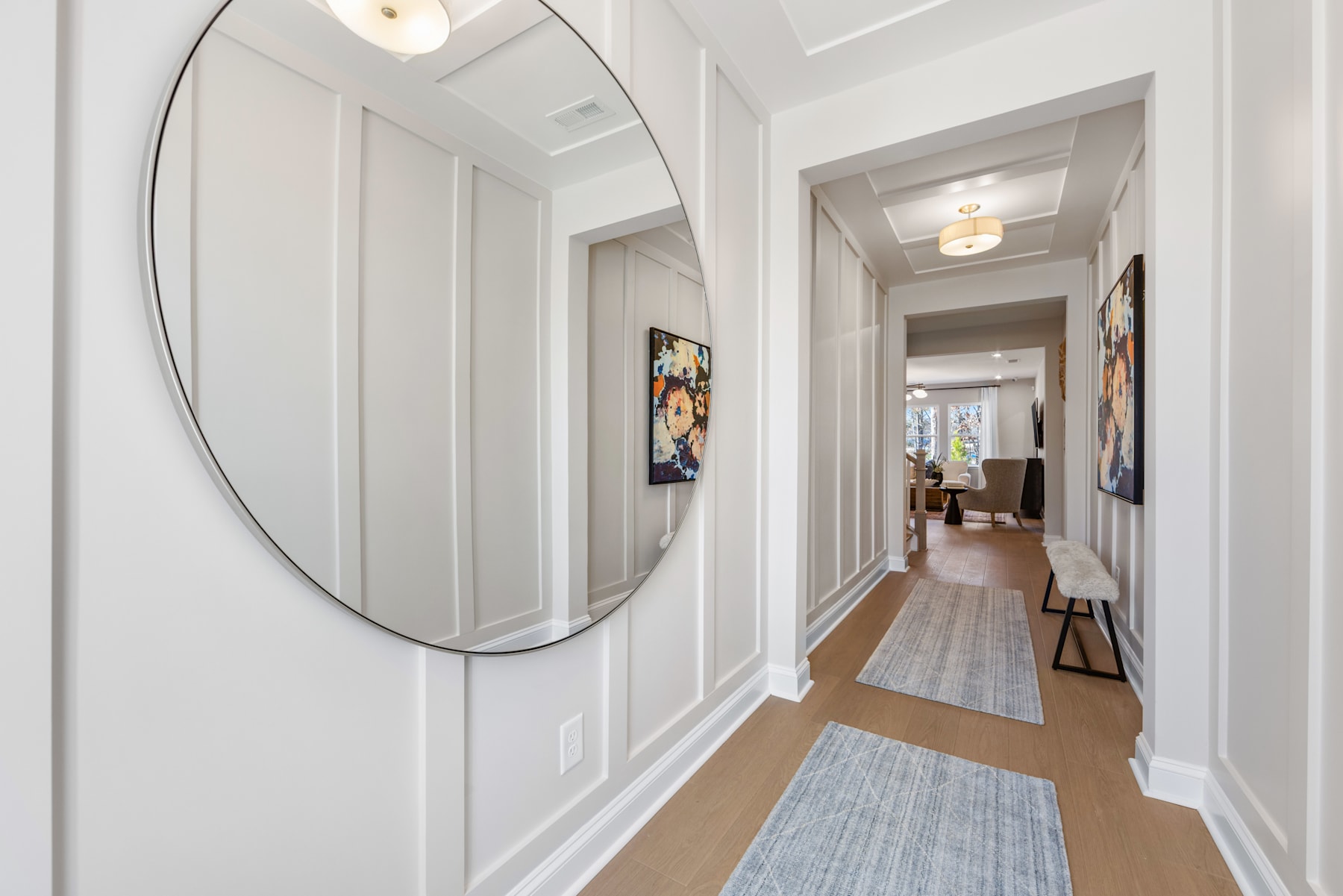 A spacious and well-lit hallway with a large oval mirror, artwork on the walls, and a wooden floor leading to a room in the background.