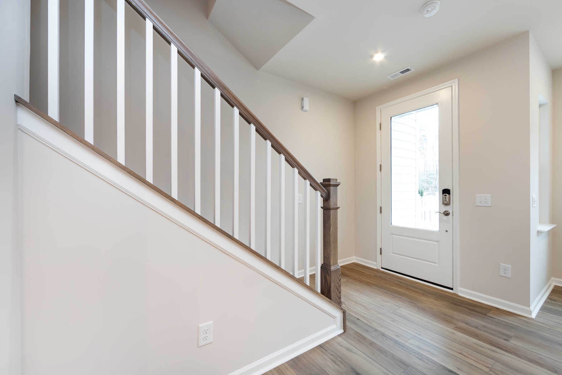 A bright and airy entryway with a wooden staircase and railing, leading to a white door that provides a glimpse of the exterior.