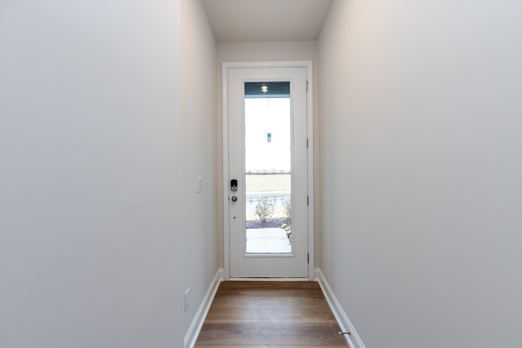 A narrow hallway with a white door leading to the outside, featuring a wooden floor and plain white walls.