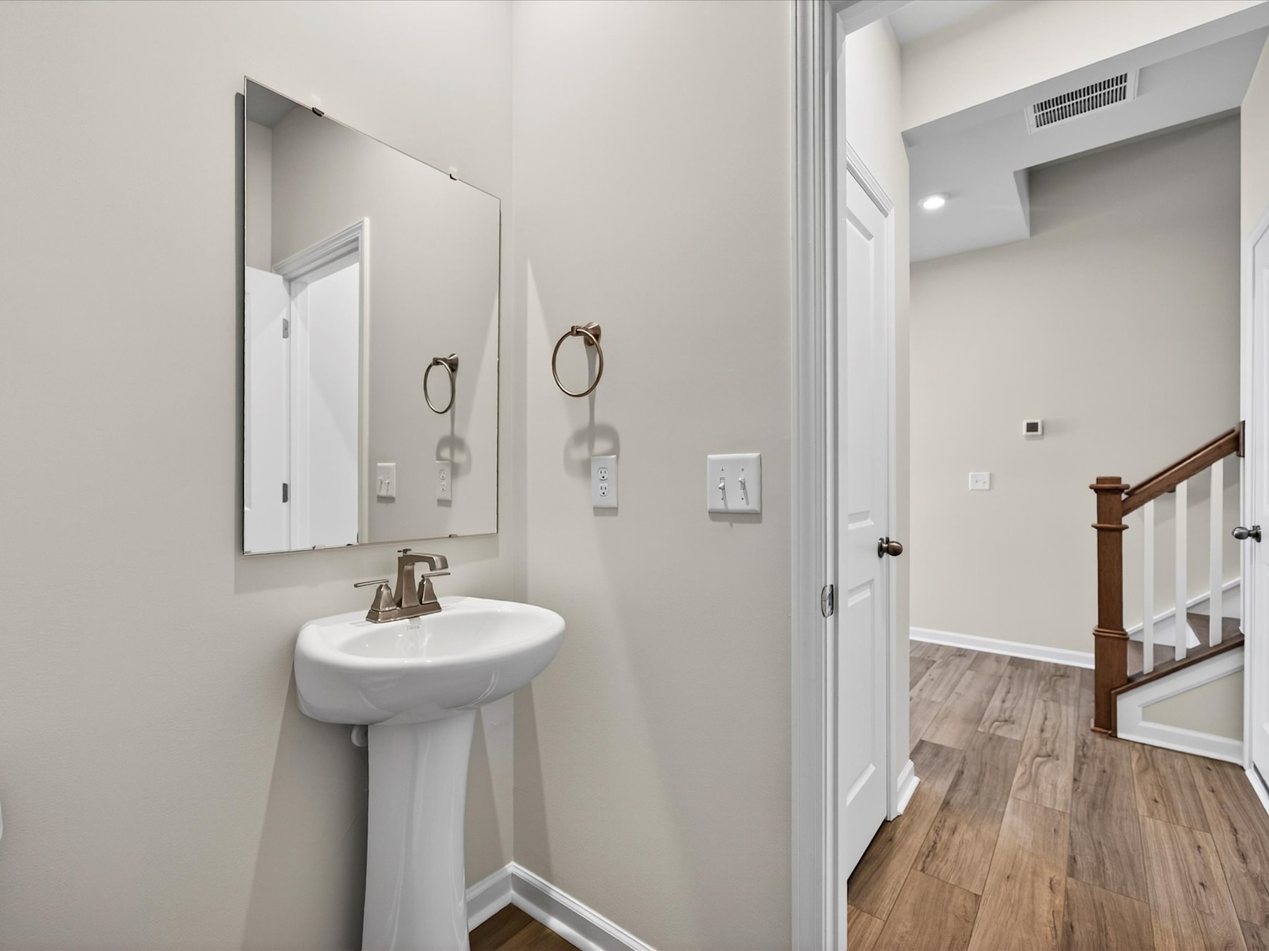 A clean, modern bathroom with a pedestal sink, mirrors, and a wooden floor leading to a hallway.