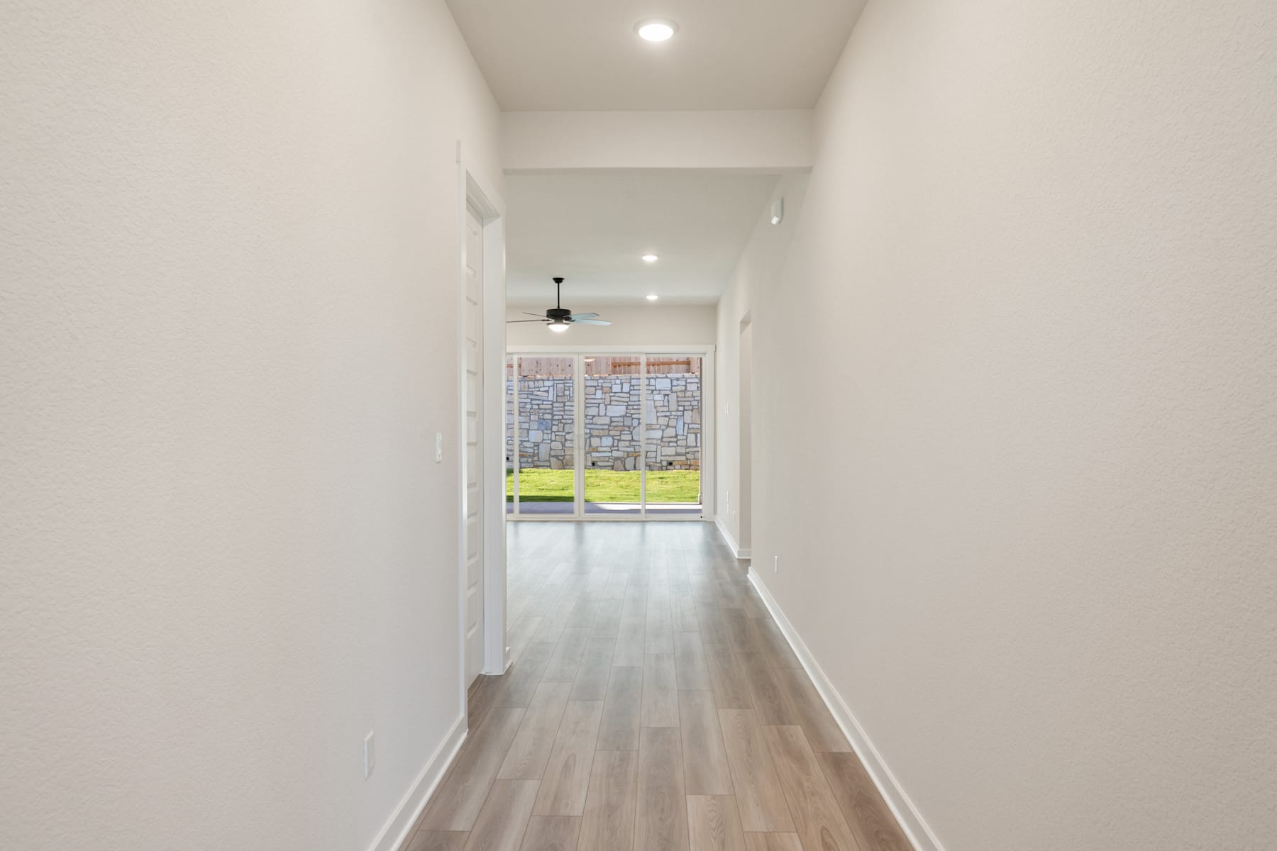 A long, well-lit hallway with white walls and a wooden floor, leading to a window at the end that provides natural light and a view of the outdoors.