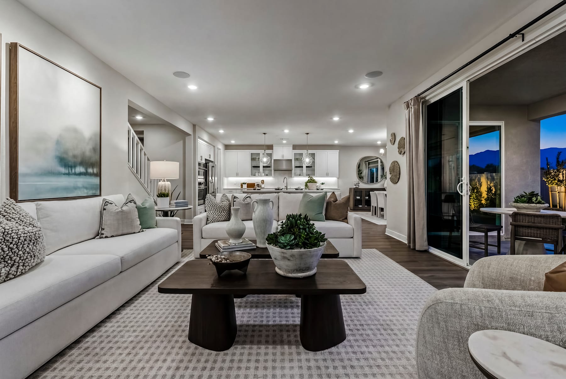 A spacious and modern living room with a cozy seating area, a large coffee table, and a kitchen visible in the background, all decorated in a neutral color palette with natural elements.