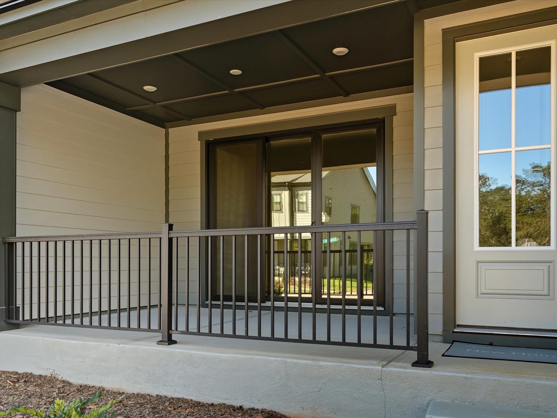 A covered porch with a metal railing leads to a wooden-framed house with large windows, surrounded by a grassy area and trees in the background.