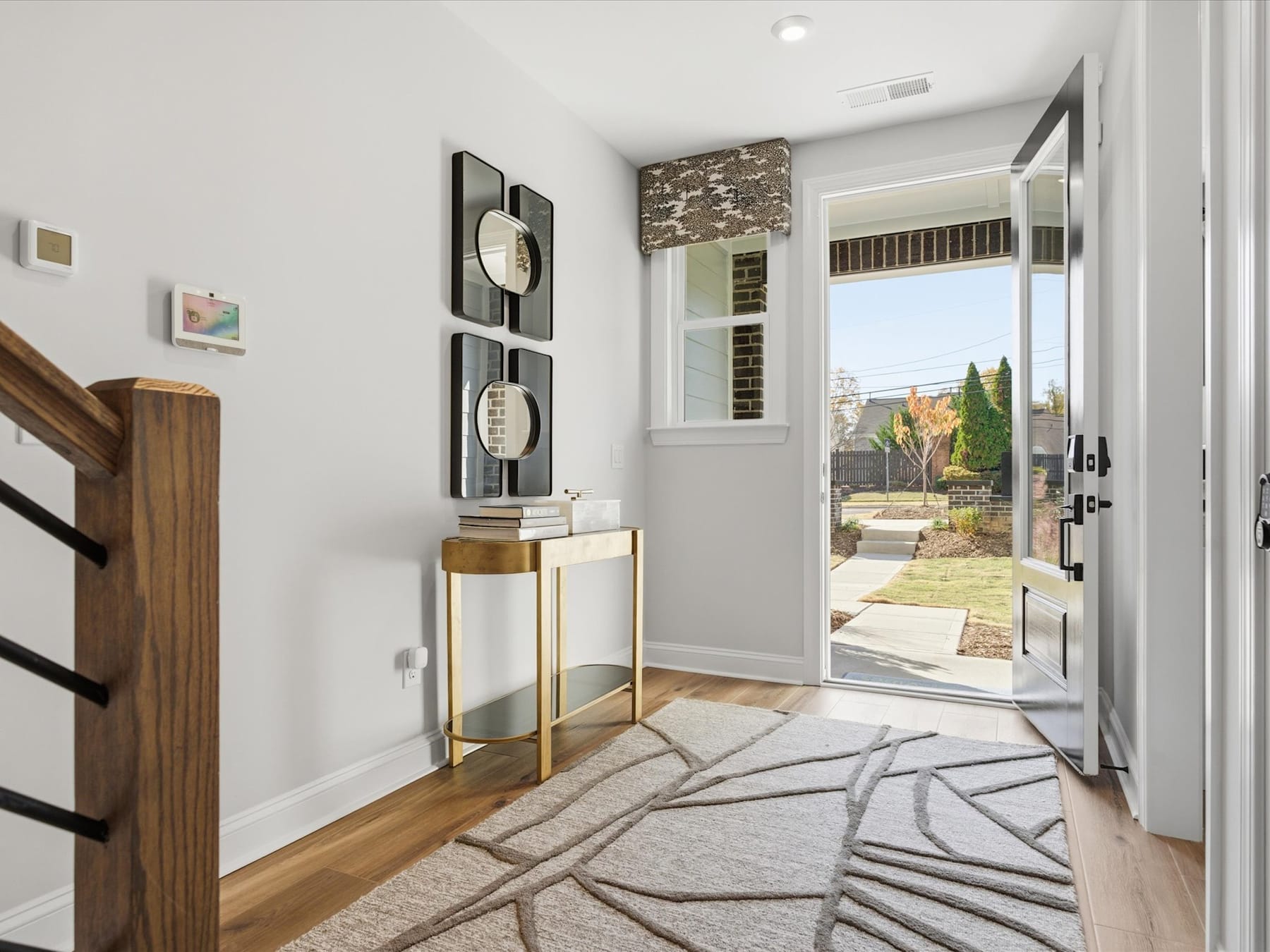 A modern and well-designed entryway with a wooden staircase, a patterned rug, and decorative wall accents, leading to a view of the outdoor landscape through a glass door.