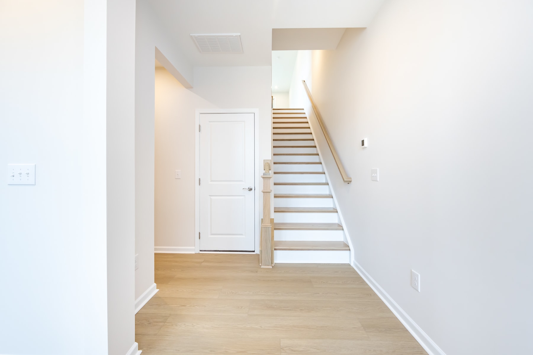 A bright and airy hallway with a wooden staircase leading upstairs, and a white door visible in the background.