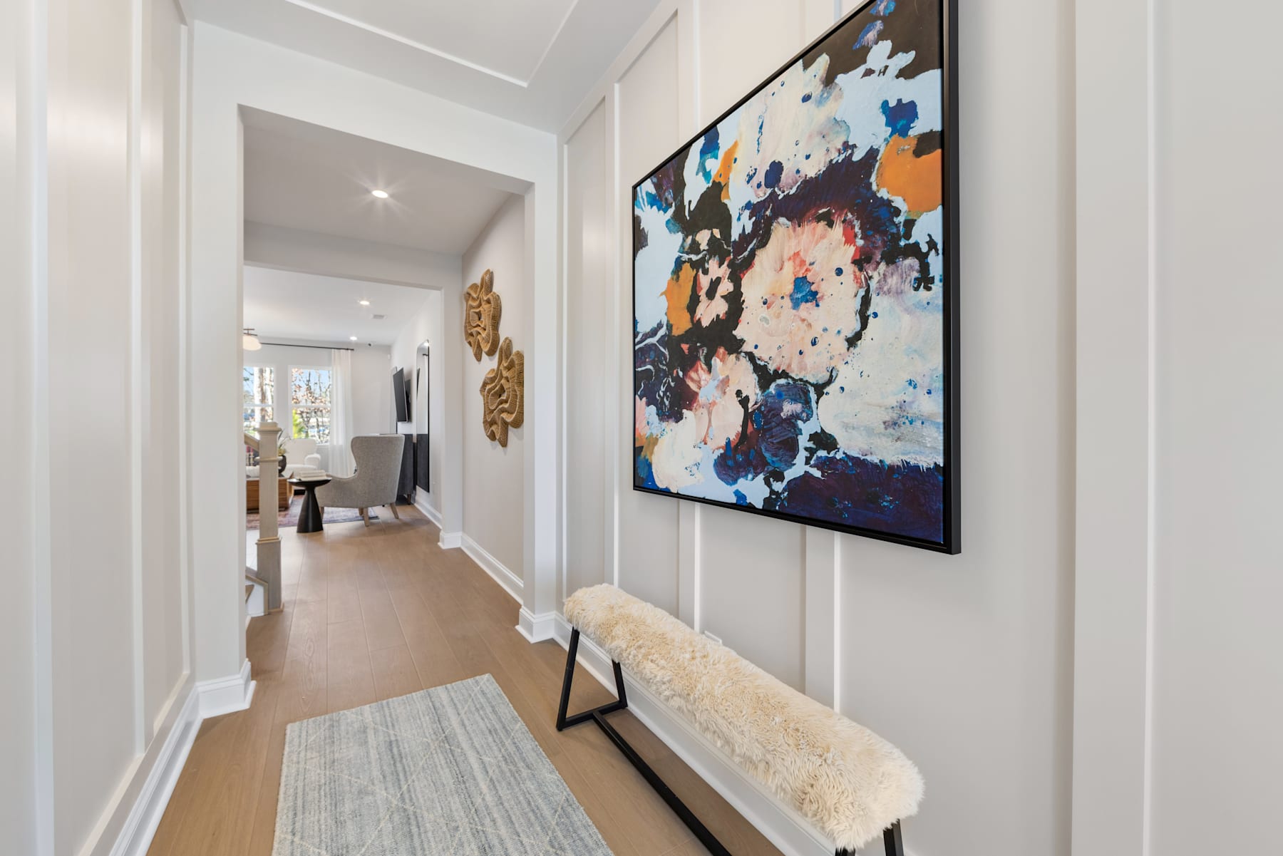 A bright and airy hallway with a large abstract painting on the wall, a white furry bench, and a wooden floor leading to a doorway in the background.