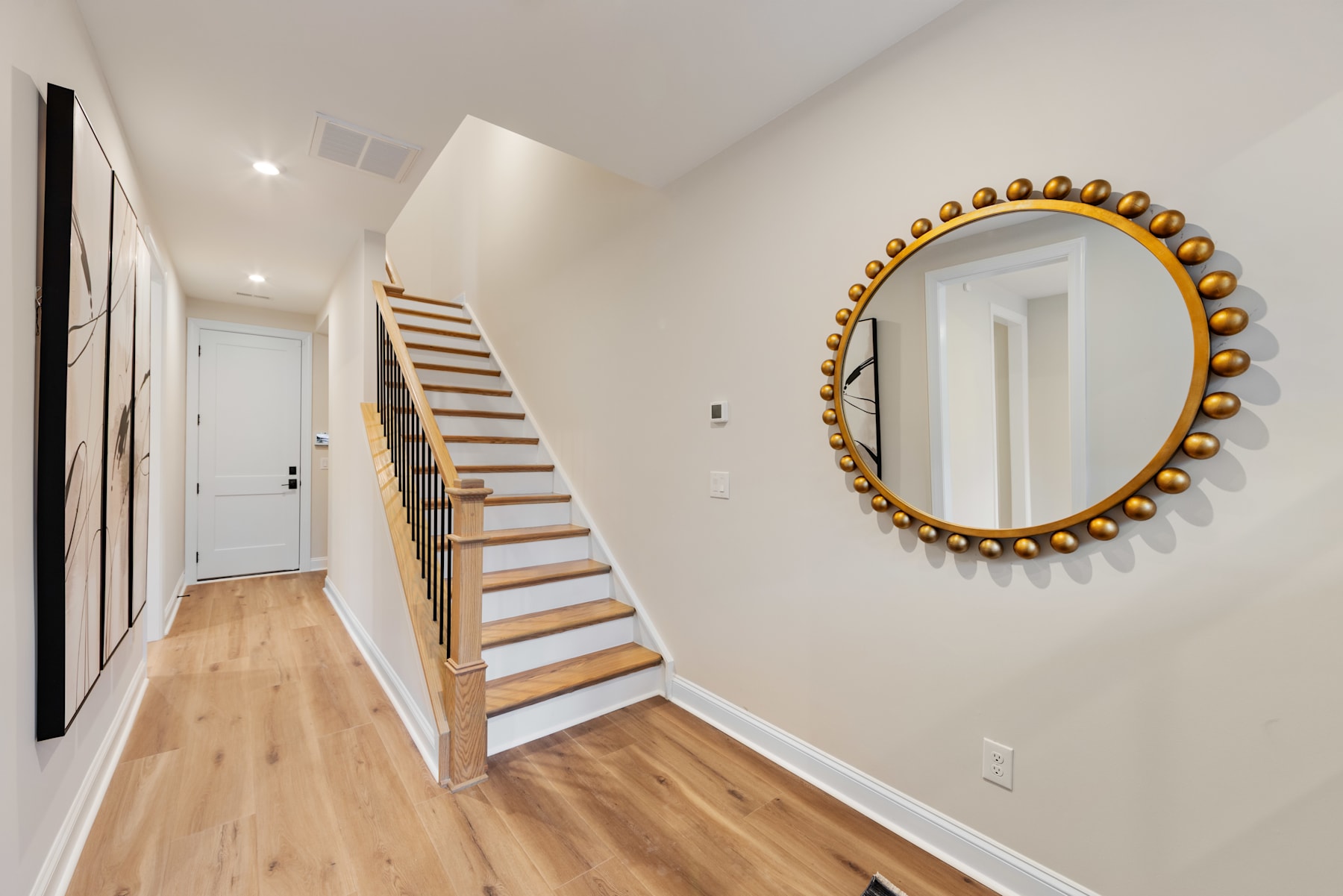 A bright and airy hallway with a wooden staircase, a large circular mirror with a golden frame, and a hardwood floor.