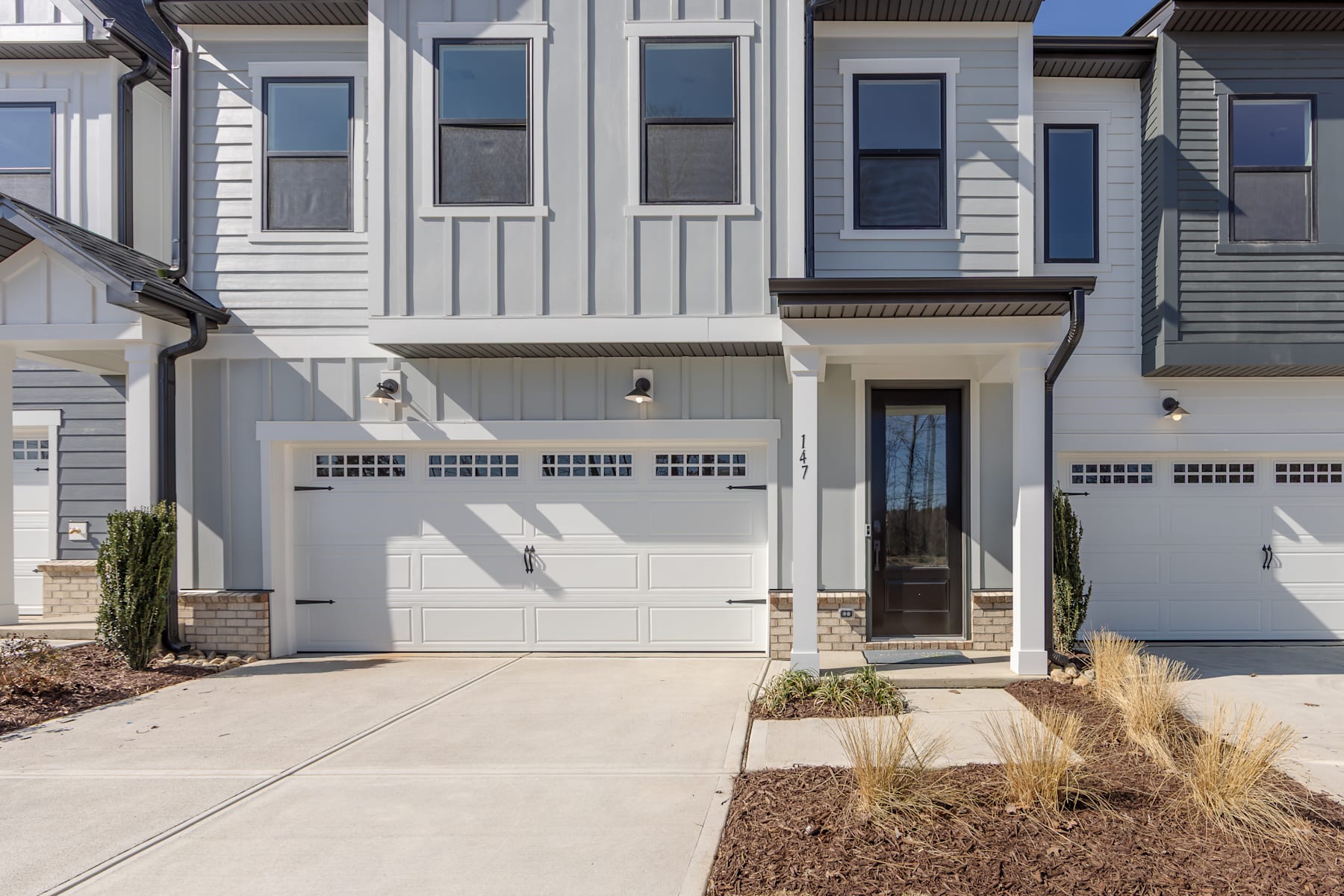 A modern, two-story townhouse with a gray exterior, white garage doors, and a paved walkway leading to the front entrance.